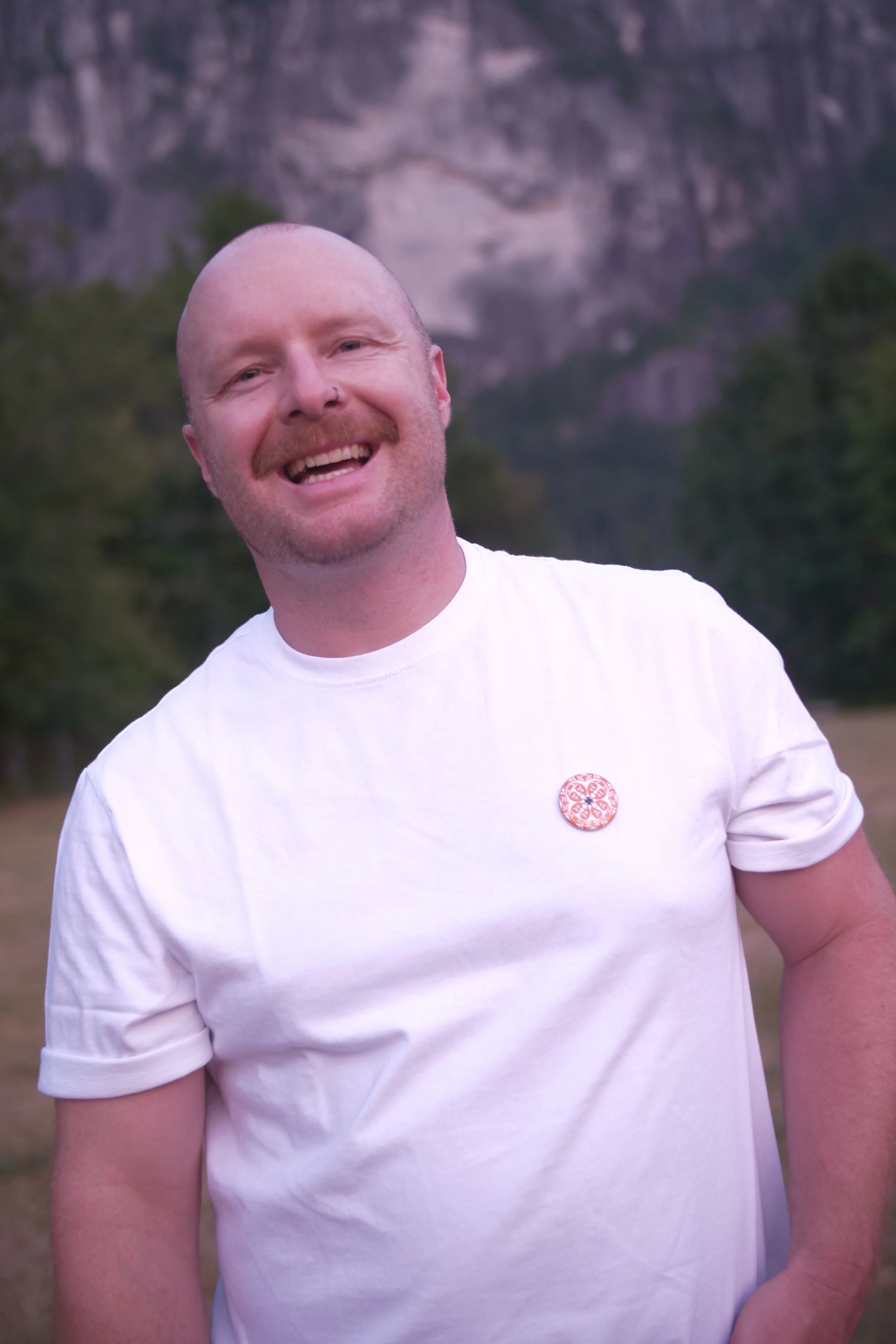 Sean - a smiling man with a bald head, mustache, and nose piercing - is wearing a white t-shirt with a floral button on the chest. He's smiling directly at the camera and looks relaxed, standing in front of the Squamish Chief mountain.