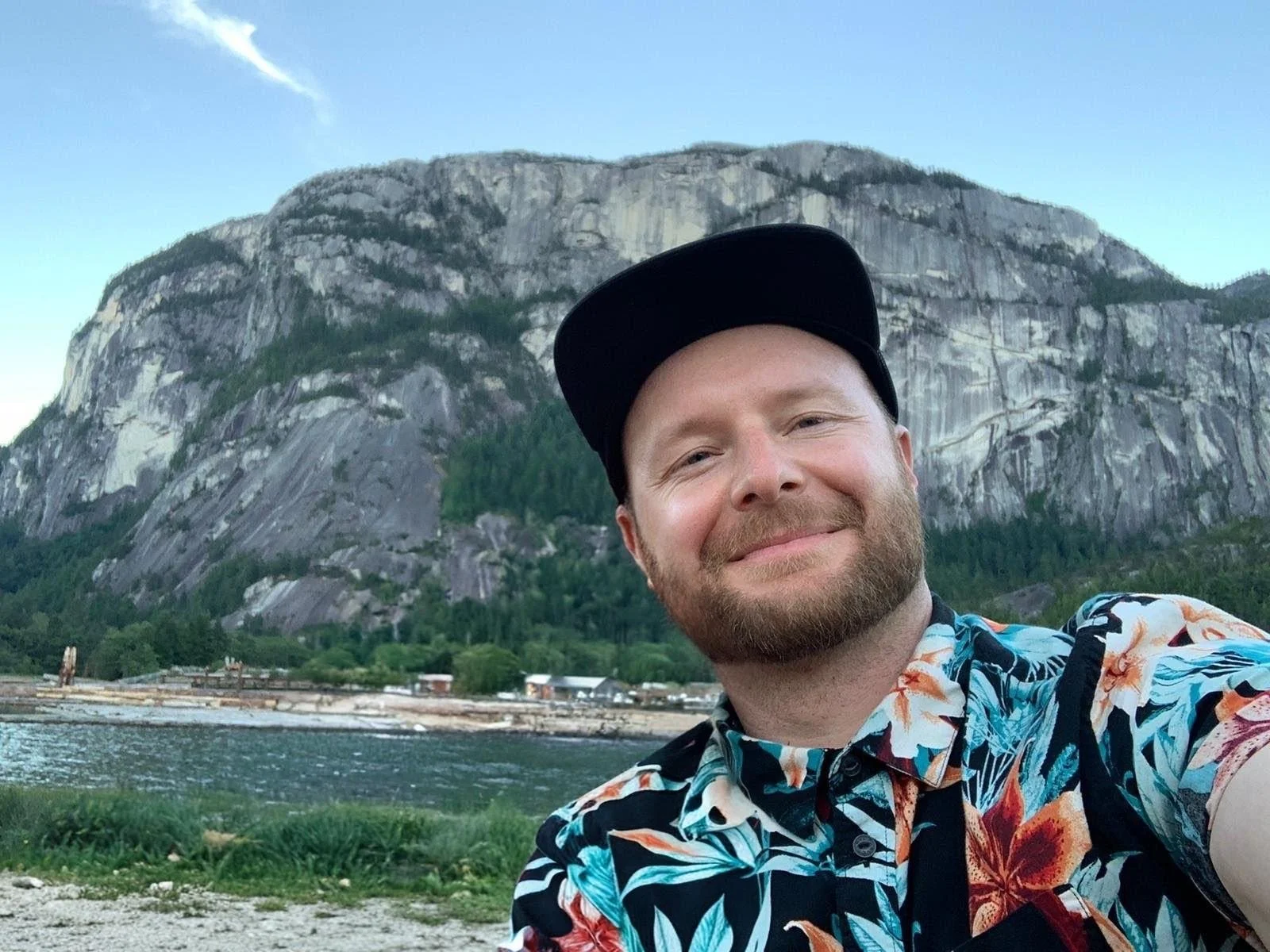 Sean Easton - a man in his 30s with a beard and a floral shirt - is taking a selfie in front of the Squamish Chief mountain, with the Mamquam channel in the mid-ground.