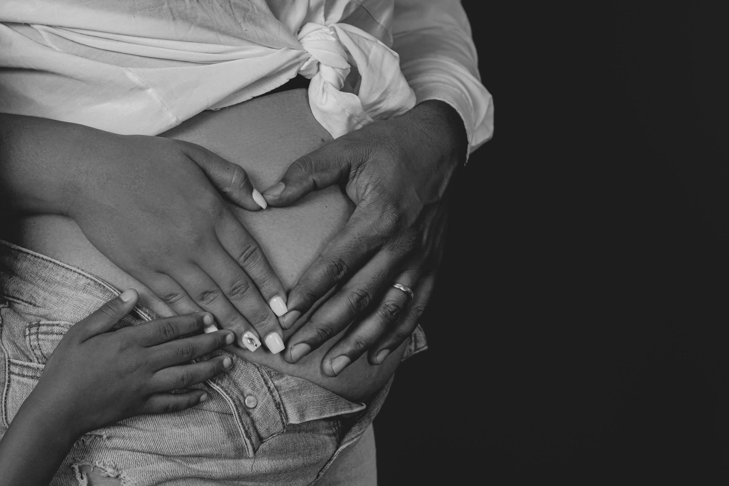 Close-up of multiple hands of different skin tones forming a heart shape on a person's midsection, with a monochrome background.