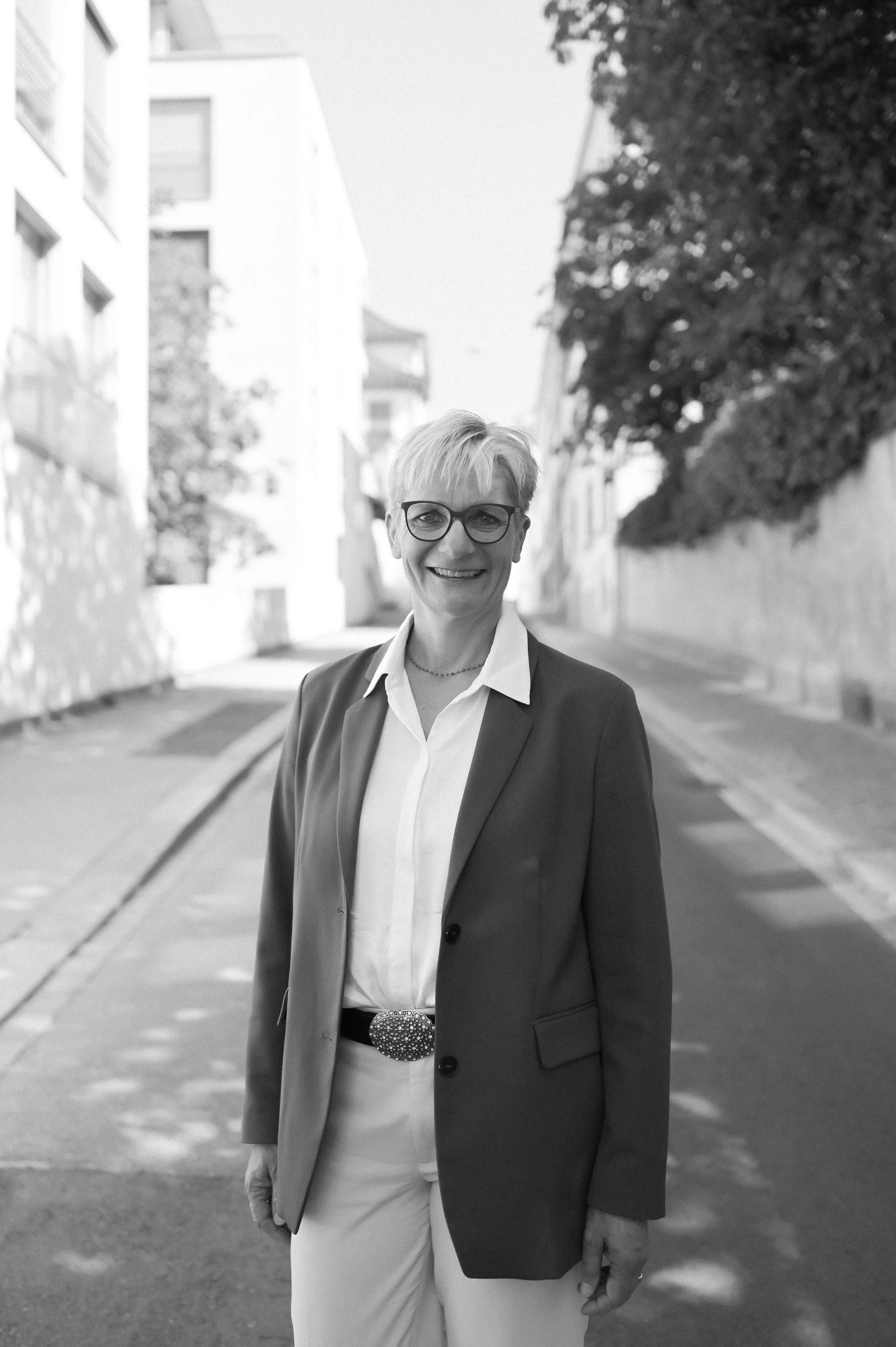 Eine Frau mit Kurzhaarfrisur und Brille, trägt einen Anzug, steht auf einer Straße mit Gebäuden im Hintergrund, lächelt, fotografiert in Schwarz-Weiß.