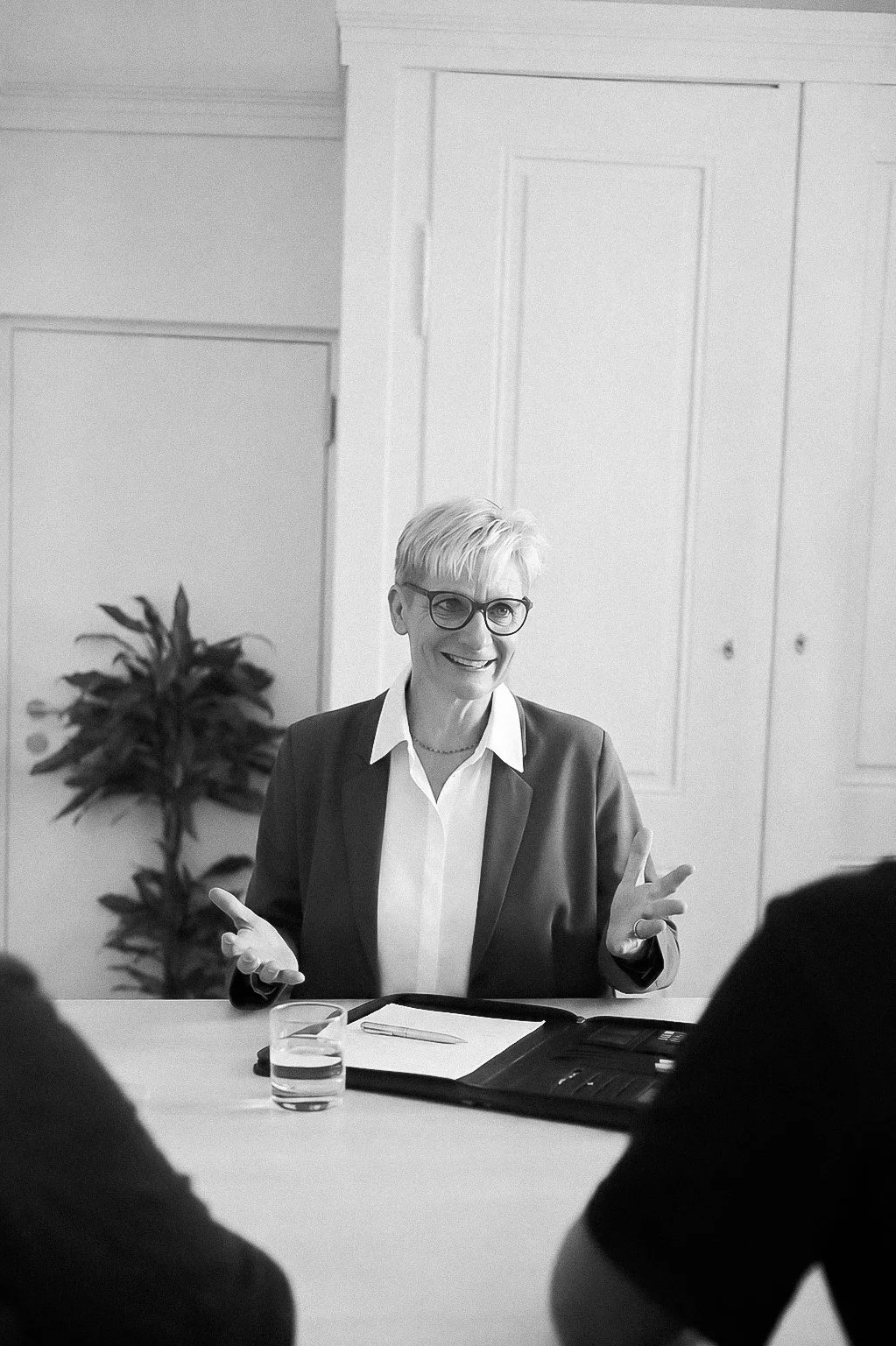 Eine Frau sitzt an einem Tisch, trägt eine Brille und eine dunkle Jacke, spricht und gestikuliert während eines Gesprächs, im Hintergrund eine Pflanze und eine weiße Tür.