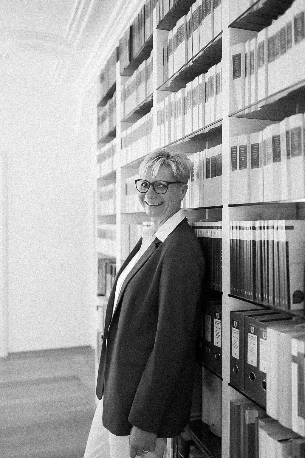 A woman with short hair, glasses, and a bright smile, wearing a blazer and white shirt, leaning against a bookshelf filled with binders and books in a library or office.