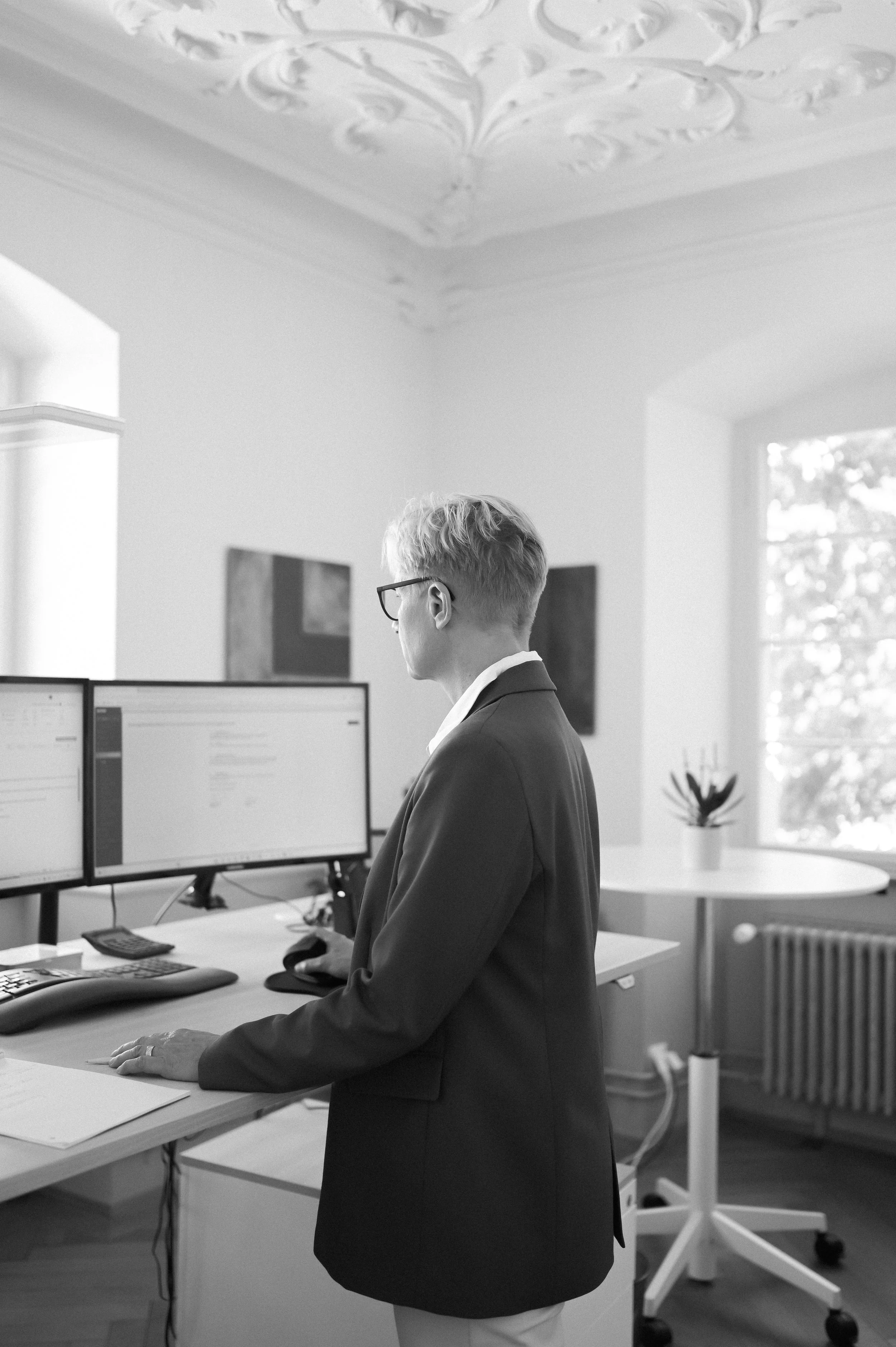 Ein Mann steht an einem Schreibtisch in einem Büro, betrachtet zwei Computerbildschirme. Er trägt eine dunkle Jacke und Brille, im Hintergrund sind Fenster und Wanddekorationen sichtbar.