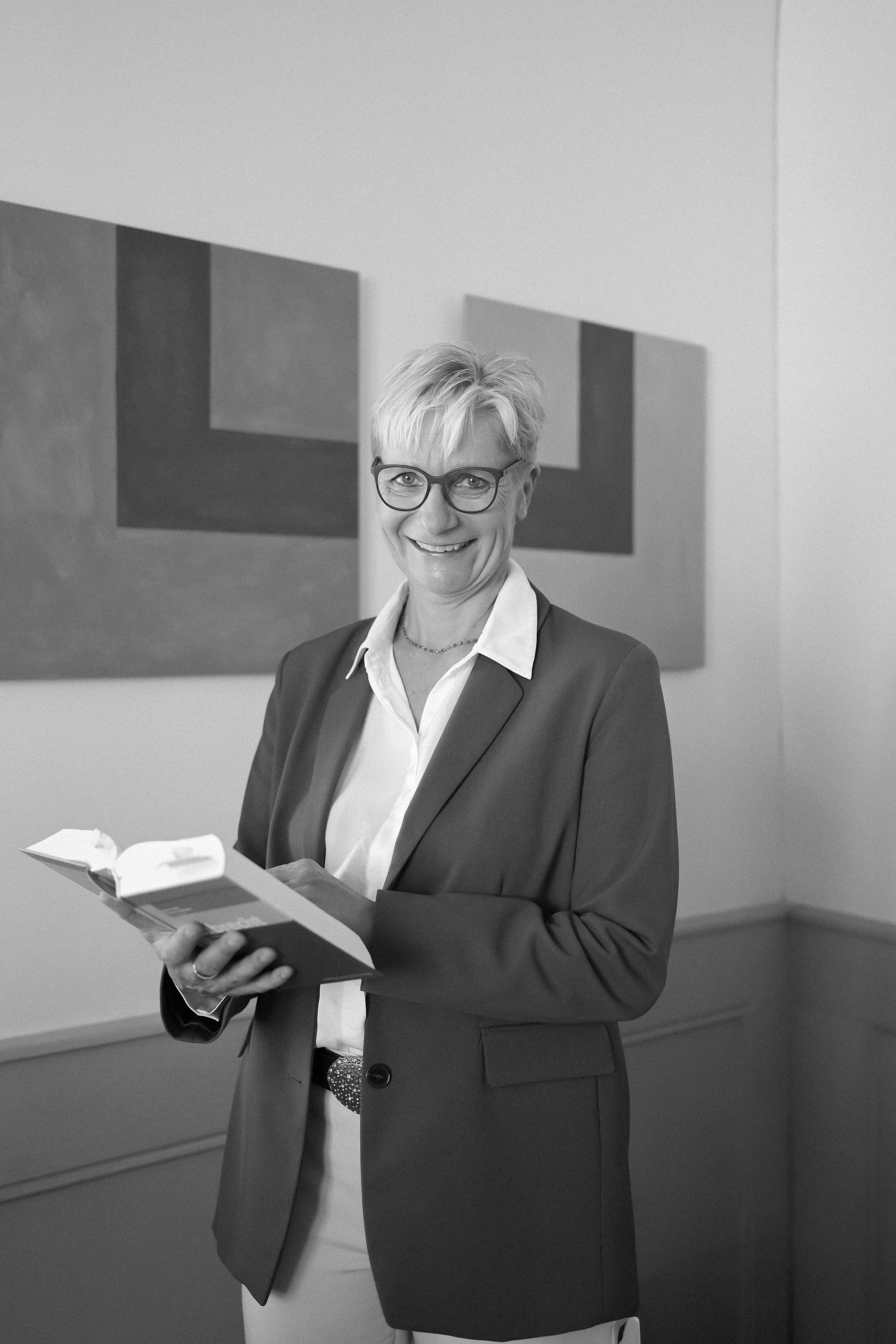 Eine lachende Frau in einem schwarzen Blazer und Brille liest in einem Buch. Im Hintergrund befinden sich geometrische Wandbilder.