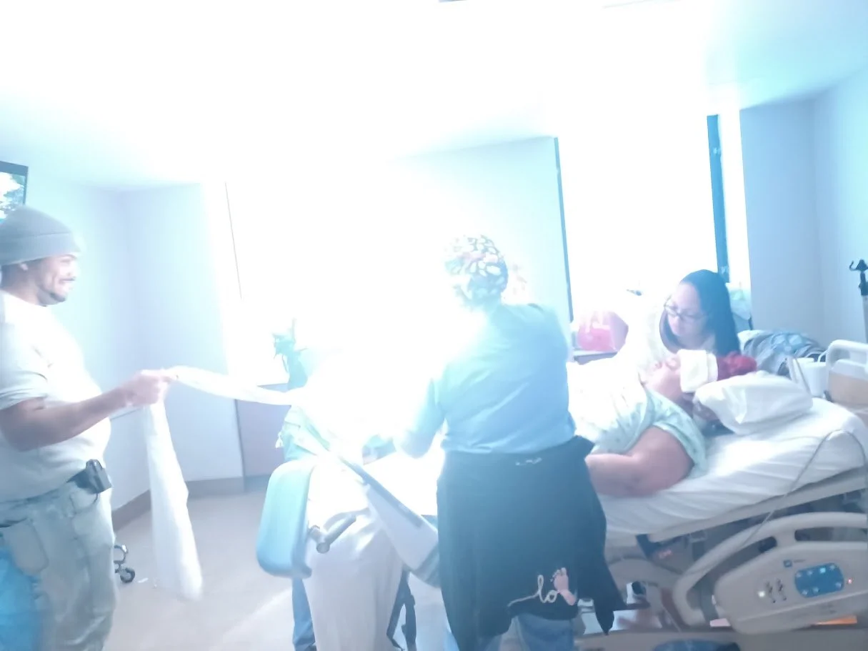 A woman in hospital bed with her newborn baby, surrounded by two women and a man with a gift, in a hospital room.