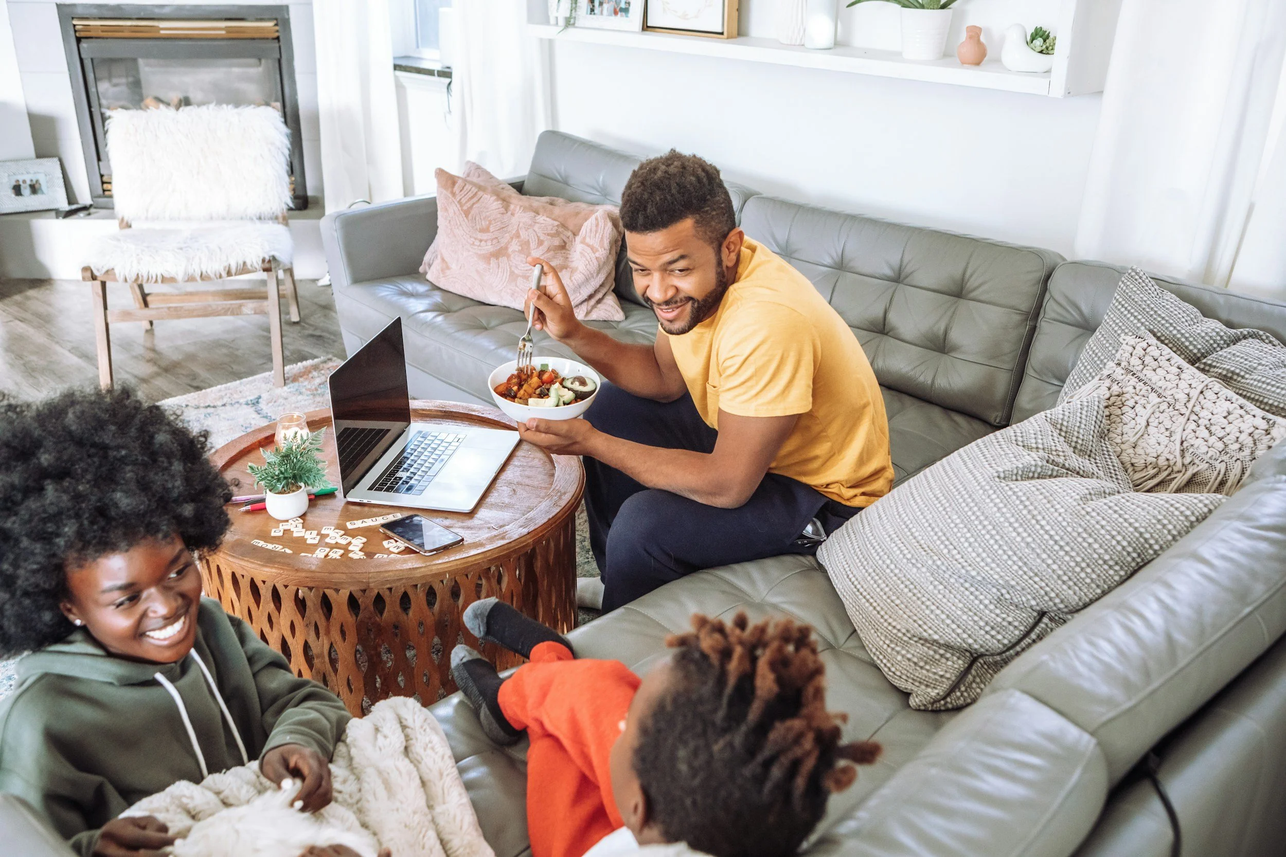 A group of people gathered in a cozy living room, sitting on a gray couch and a wooden chair, enjoying a meal and conversation. There is a laptop on the coffee table, and the room is decorated with plants, framed pictures, and pillows.