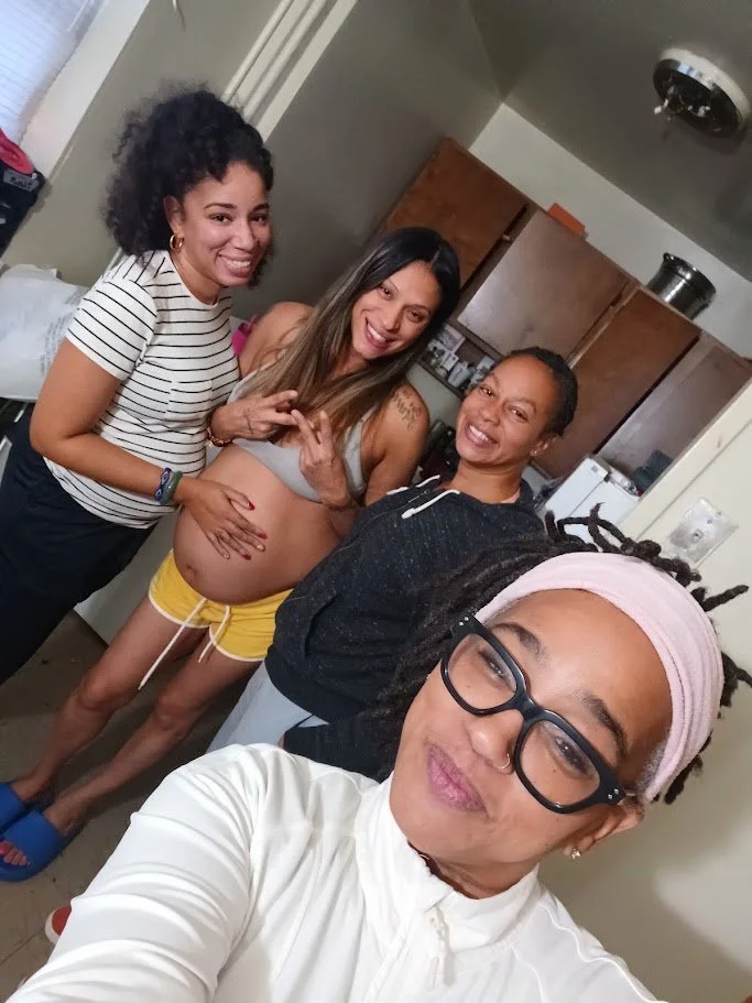 Four women are posing for a selfie in a kitchen, smiling and making playful gestures. One woman is pregnant, wearing a gray sports bra and yellow shorts. The others are dressed casually, with one wearing a striped shirt and black pants, another with a hoodie, and the fourth with glasses and a headscarf.