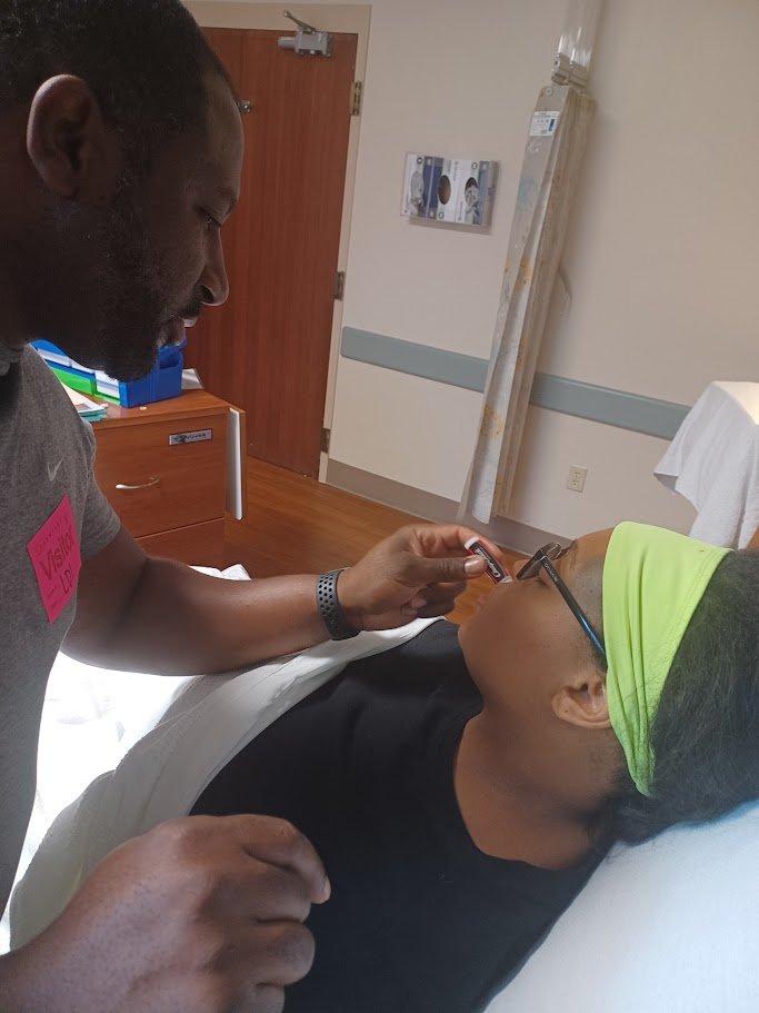 A man is administering a nasal spray to a woman lying on a hospital bed, who is wearing glasses and a neon green headscarf.