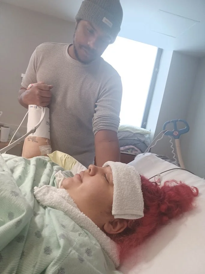 A woman with red hair lying in a hospital bed, wearing a towel on her head and a hospital gown. A man standing beside her is holding her arm, with an IV attached, and appears to be watching her.