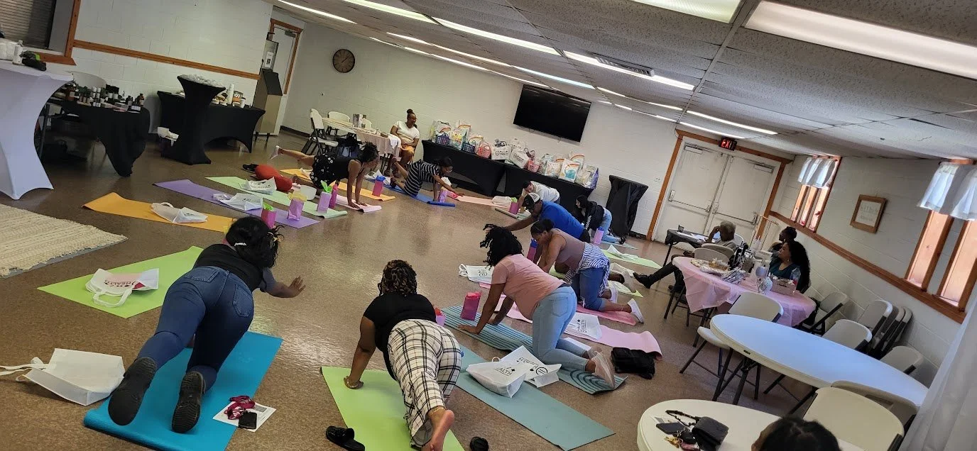 Group of women participating in a yoga class on yoga mats in a community room with tables and chairs.