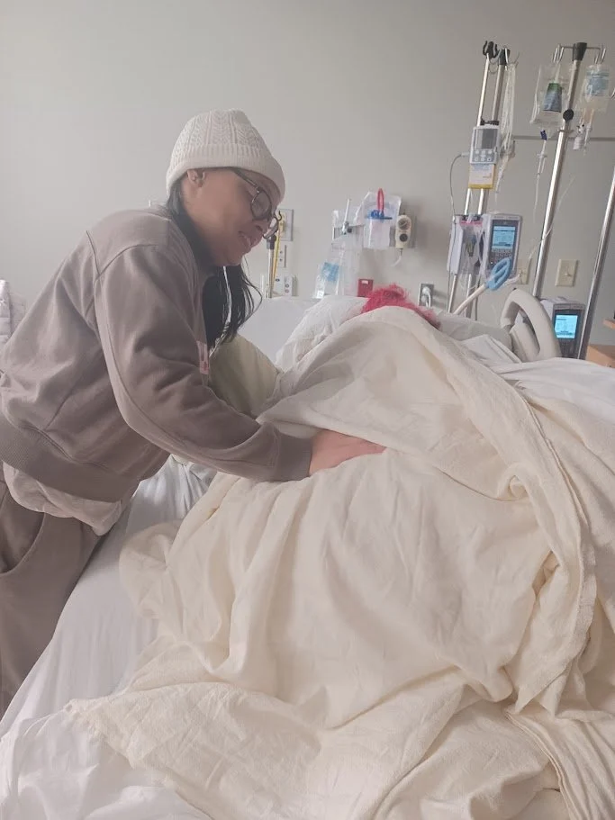 A woman wearing glasses and a knit hat is leaning over a patient in a hospital bed, gently touching their shoulder. The patient is covered with a cream-colored blanket and has bright pink hair, lying on the hospital bed. Medical equipment and monitors surround the bed in a clinical setting.