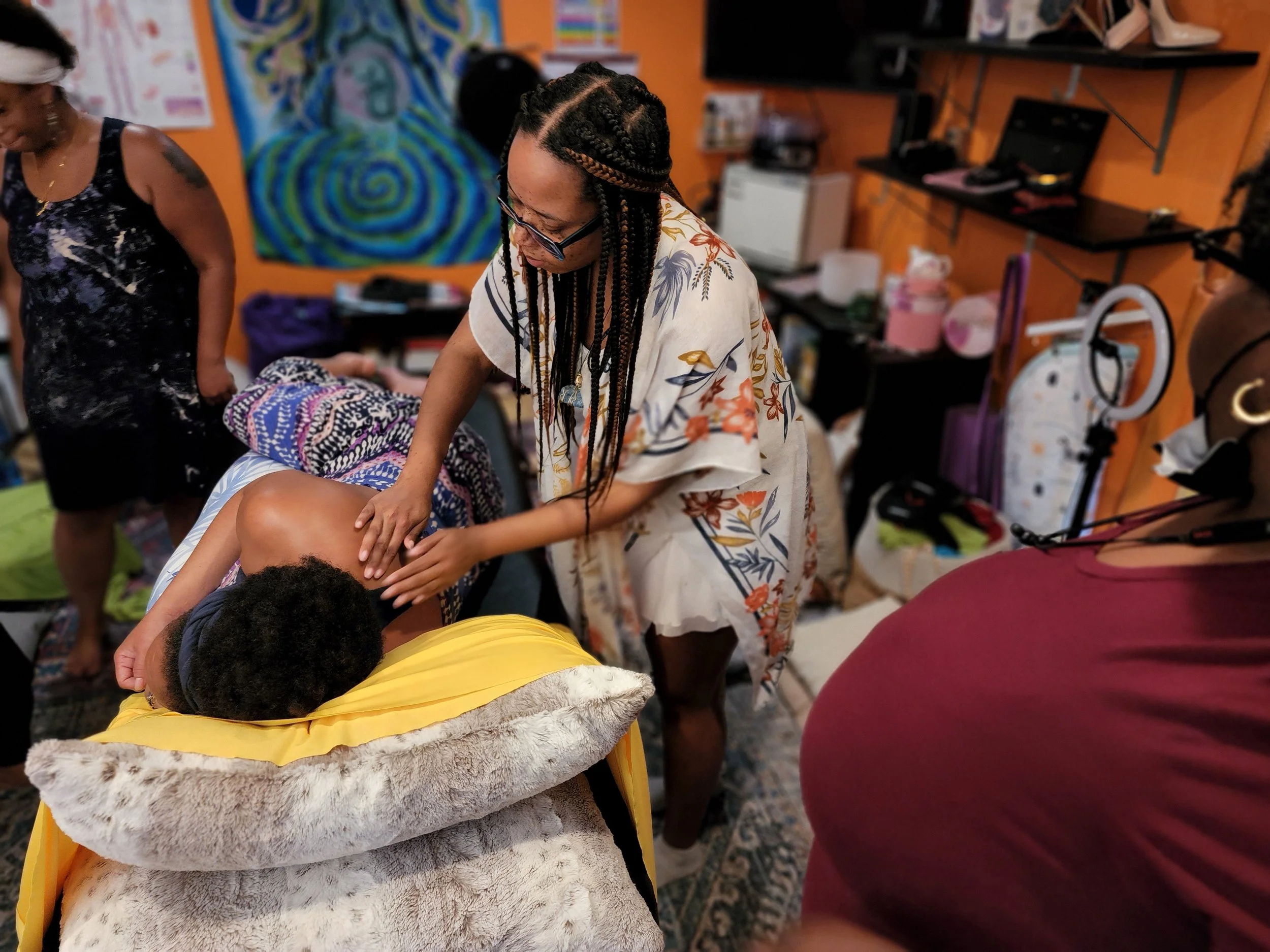 Woman with braided hair and glasses giving a massage to a person lying face down on a bed, surrounded by other women in a colorful room.