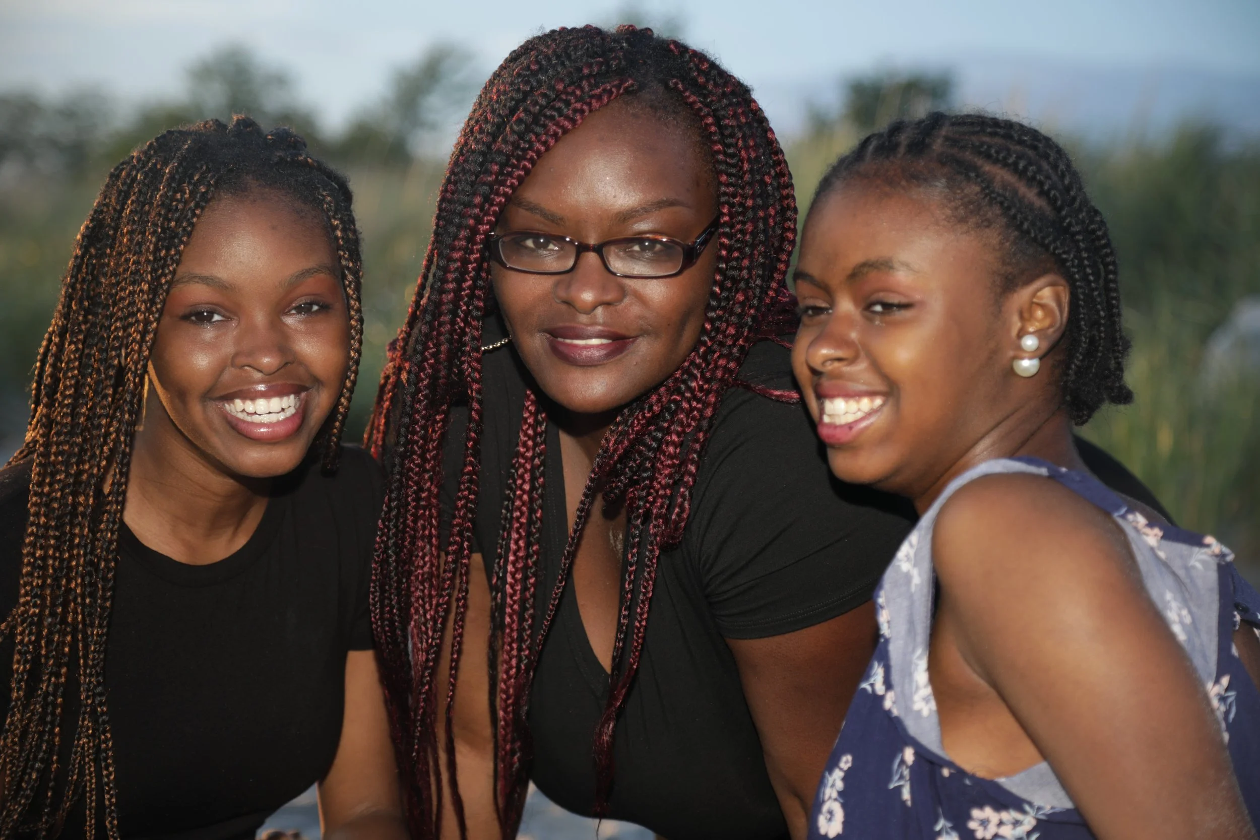 Three women with braided hair sharing a close moment outdoors, smiling and appearing joyful.