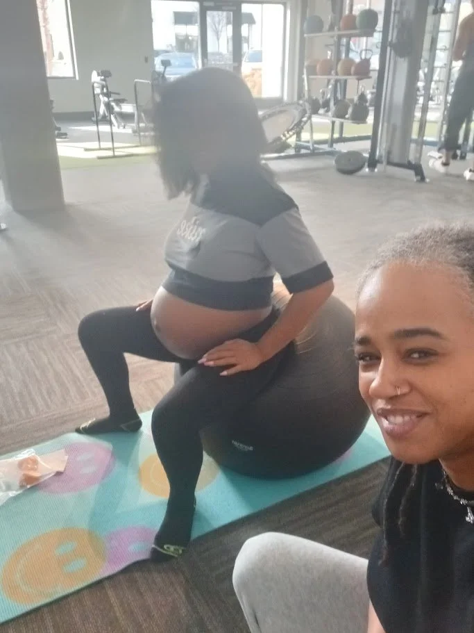 A pregnant woman in workout clothes sitting on a fitness ball at a gym, smiling at the camera. A woman in workout clothes is sitting on the fitness ball, and there are gym equipment and windows in the background.