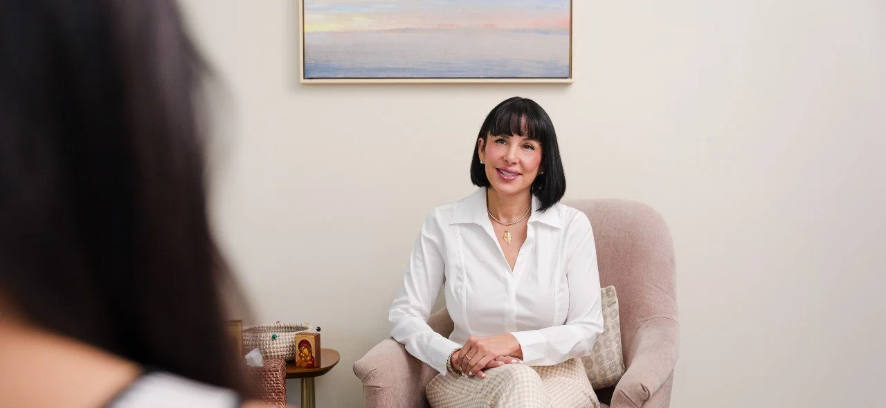 A woman with black hair and bangs sitting on a beige armchair, smiling during a conversation in a well-lit room with a artwork on the wall behind her.