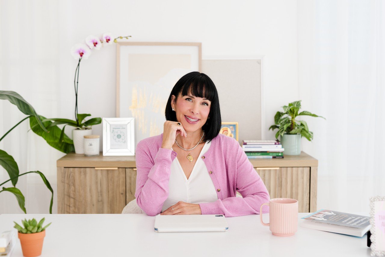 Smiling woman sitting at a desk in a bright office with plants and decorative items.