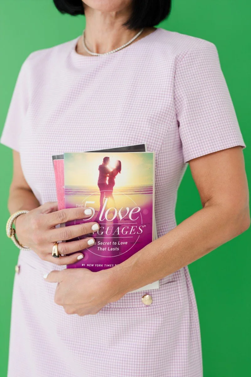 A woman in a pink checkered dress holding a book titled 'The Love Guages' against a green background.