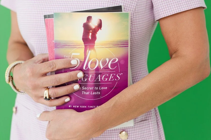 A woman holding a book titled "The 5 Love Languages: The Secret to Love That Lasts" with a silhouette of a couple on the beach at sunset on the cover.