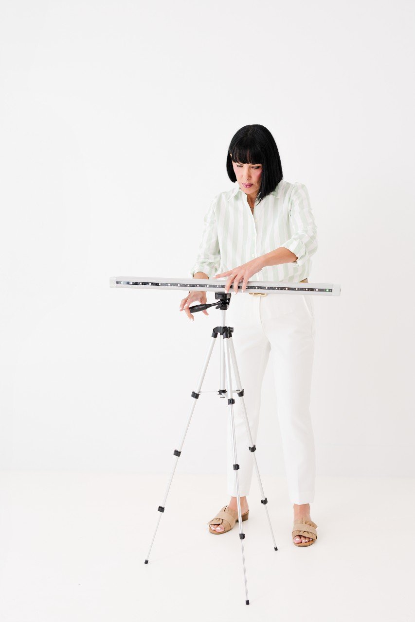 Woman standing in white outfit adjusting camera on tripod in studio with plain white background.