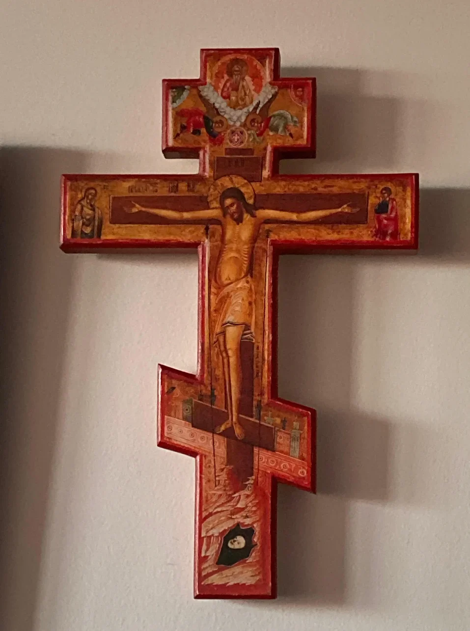 Orthodox Christian cross with colored iconography depicting Jesus Christ crucified, with saints and religious scenes, mounted on a beige wall.