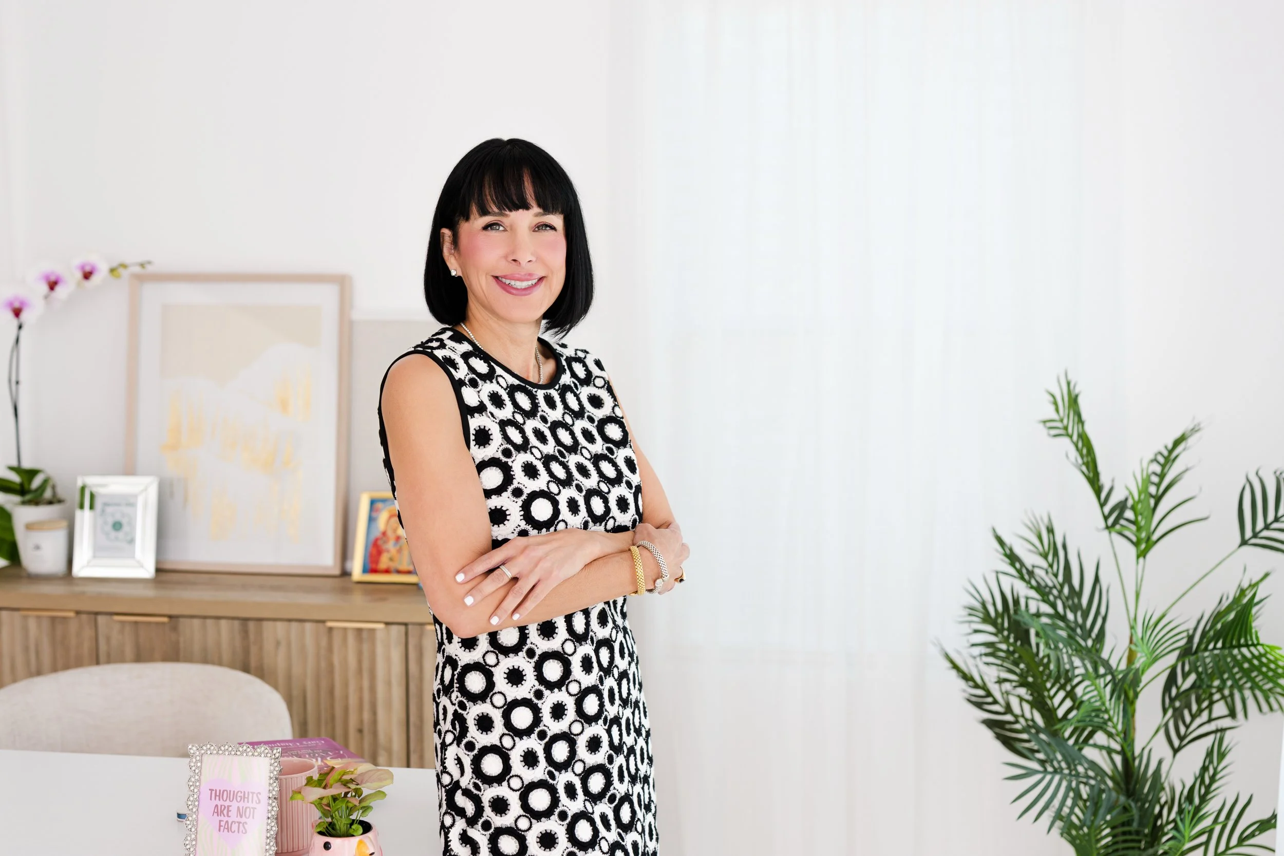 A smiling woman with black hair and a black-and-white patterned dress standing with arms crossed in a bright room with plants and framed pictures on a wooden sideboard.