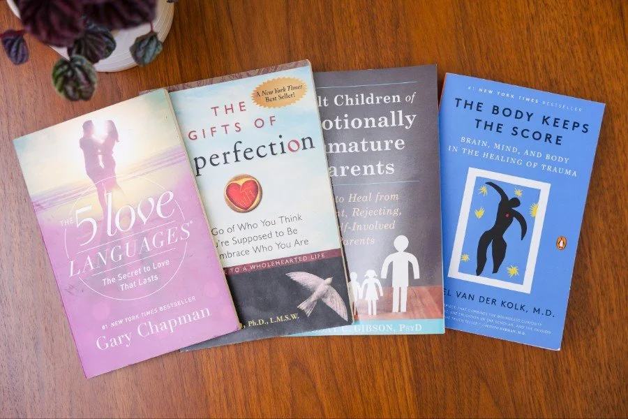 Four paperback books lying on a wooden surface with a potted plant partially visible in the upper left corner.