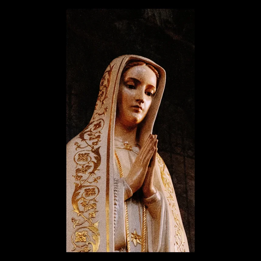 A religious statue of the Virgin Mary with hands in prayer, wearing a beige cloak with gold embroidery.