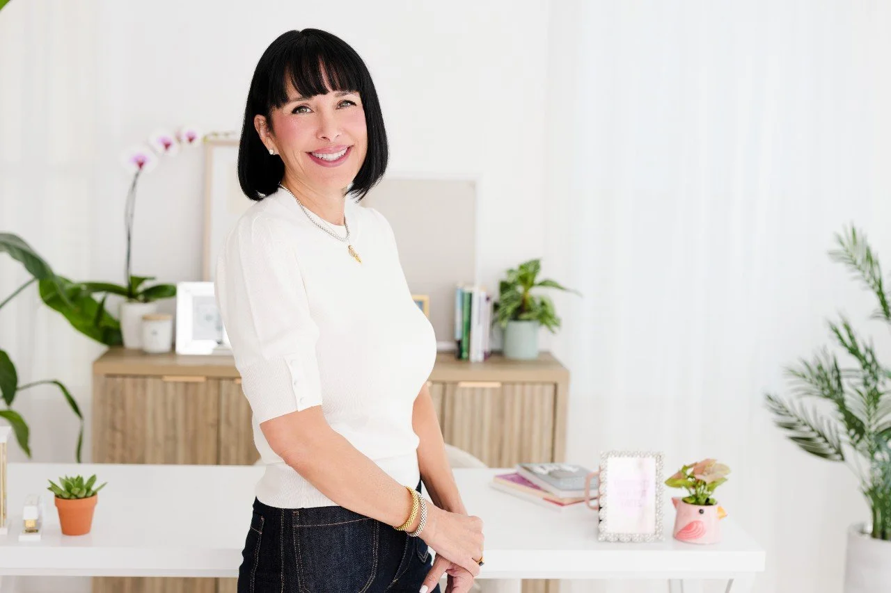 A woman with black hair, wearing a white top, standing in a modern, well-lit office or home workspace, smiling at the camera.