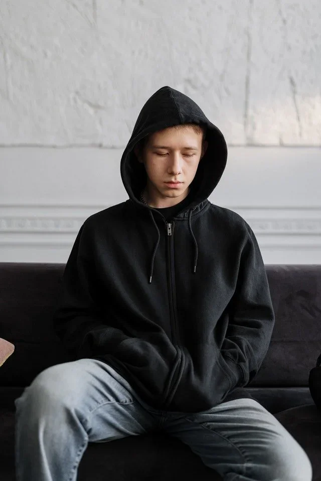 Young man in a black hoodie sitting alone on a sofa with his eyes closed, conveying emotional distress and withdrawal.