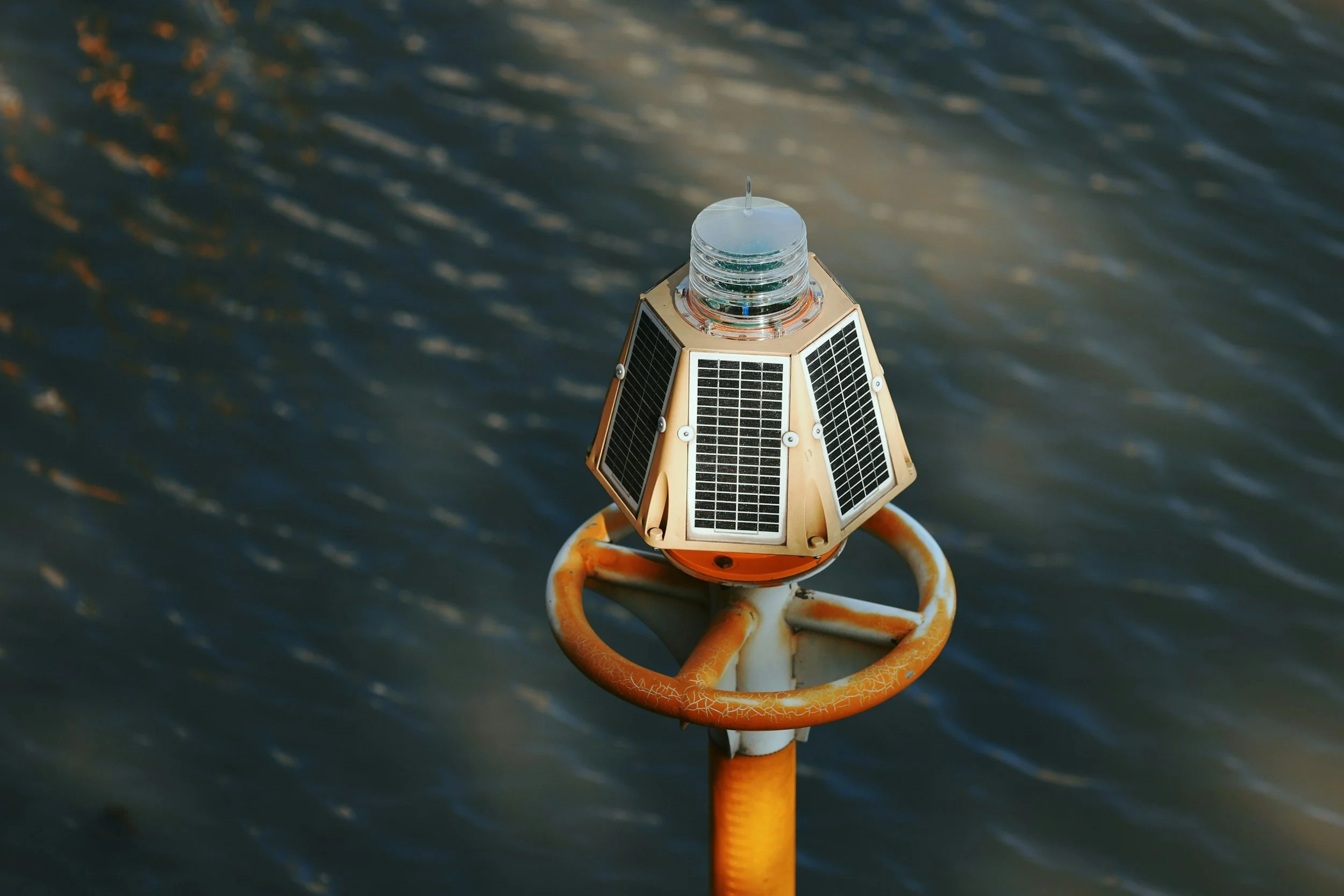 An ocean monitoring device - A small satellite or solar panel device mounted on a yellow and orange metal structure above water. The device has a transparent dome on top and several solar panels.