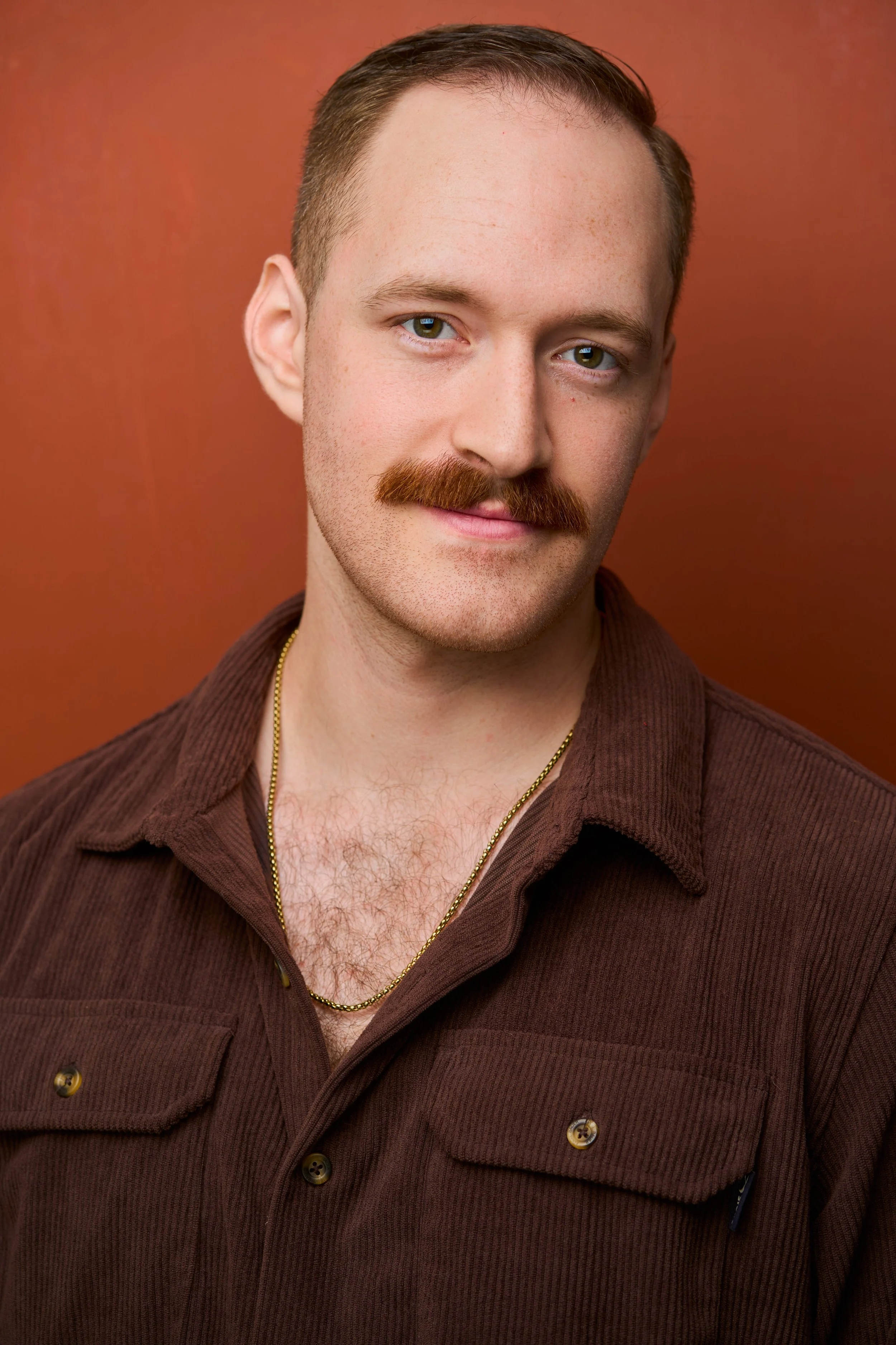 Close-up portrait of a man with a mustache, short brown hair, wearing a brown corduroy shirt and gold chain necklace, against a burnt orange background.