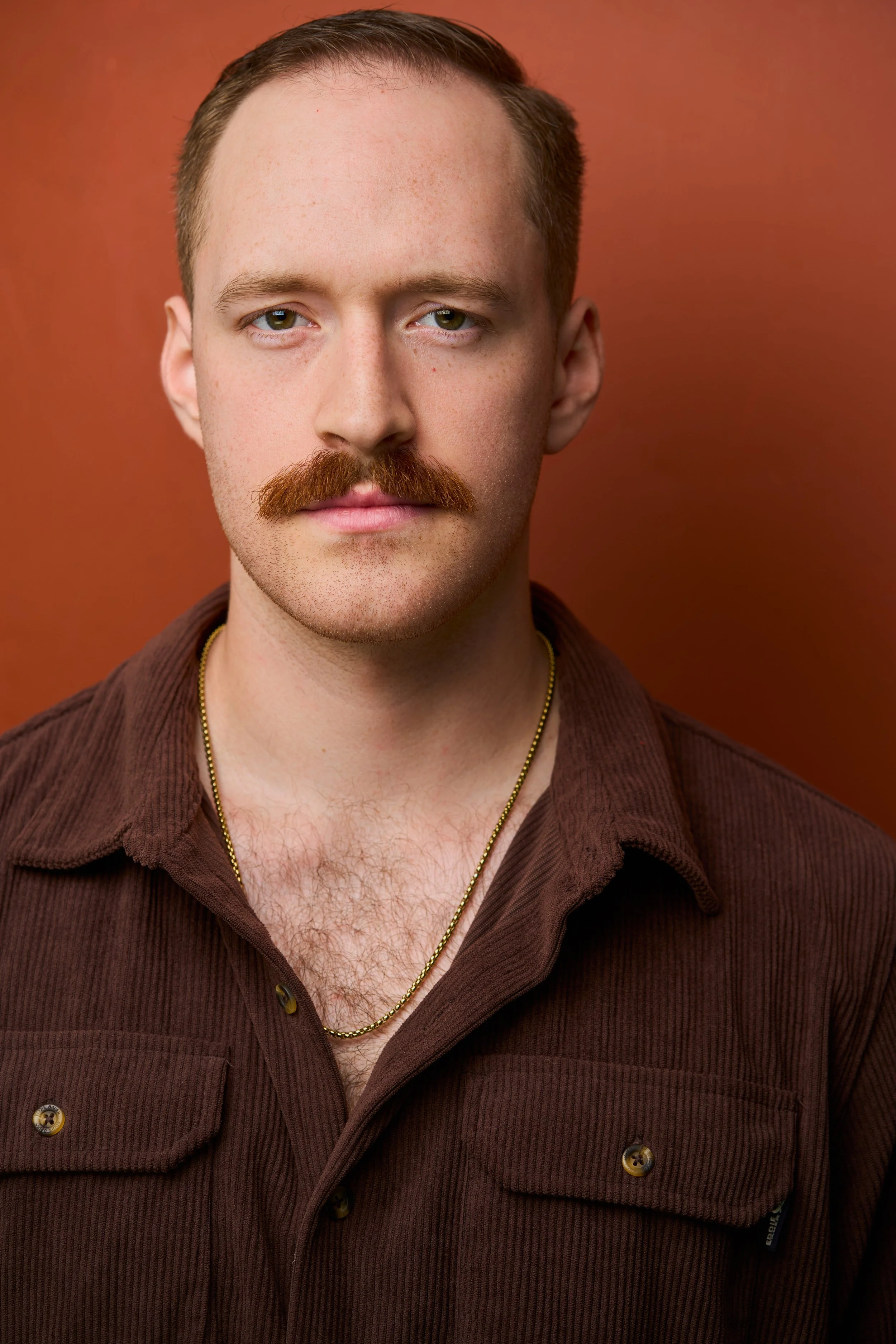 A young man with light skin, short brown hair, green eyes, and a thick mustache. He is wearing a brown corduroy jacket and a gold chain necklace. The background is a solid reddish-brown color.