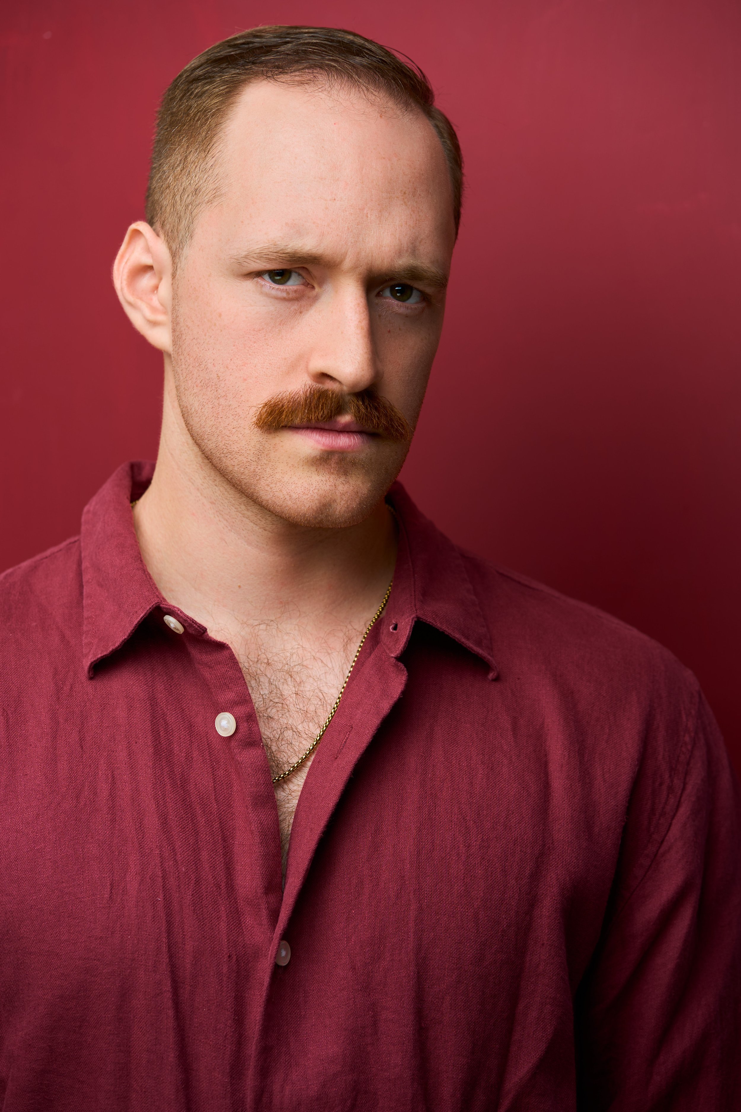 A young man with light skin, ginger hair, blue eyes, a mustache, and a beard. He is wearing a maroon button-up shirt, a gold chain necklace, and standing against a maroon background.