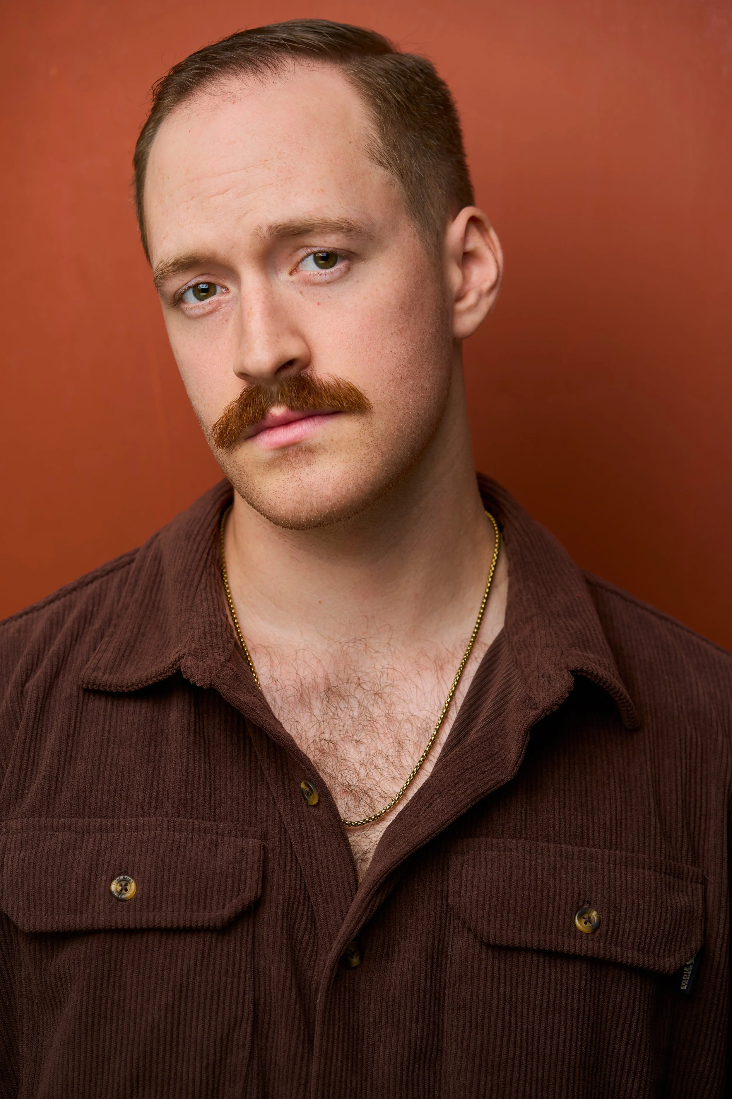 Close-up of a man with short brown hair, mustache, wearing a brown collared shirt, gold chain, posed against a reddish-orange background.
