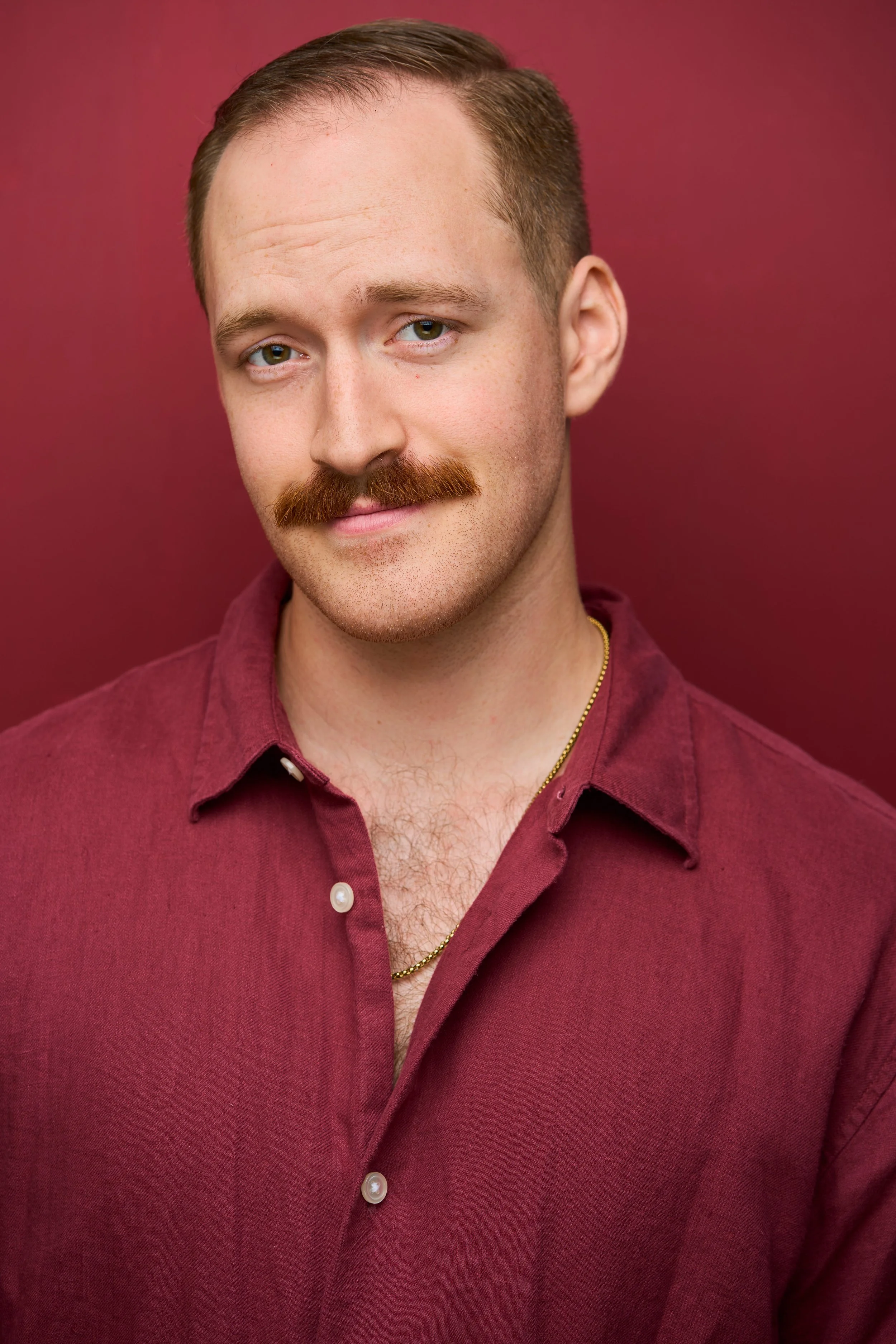 A young man with red hair, a mustache, and a slight beard, wearing a maroon shirt with the top buttons undone, standing against a solid burgundy background.