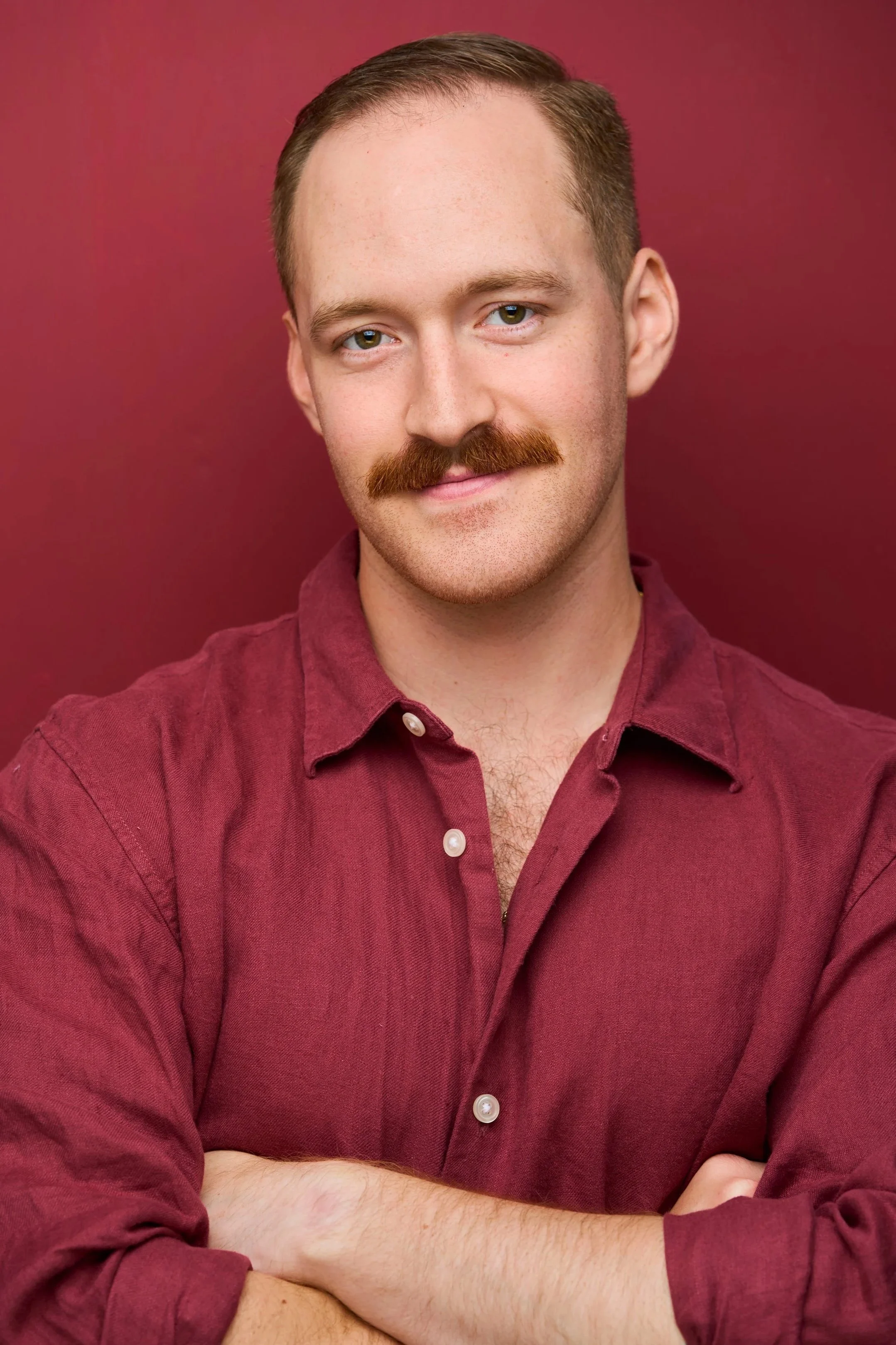 Close-up of a young man with short brown hair, hazel eyes, and a mustache, wearing a maroon button-up shirt, standing against a matching maroon background.
