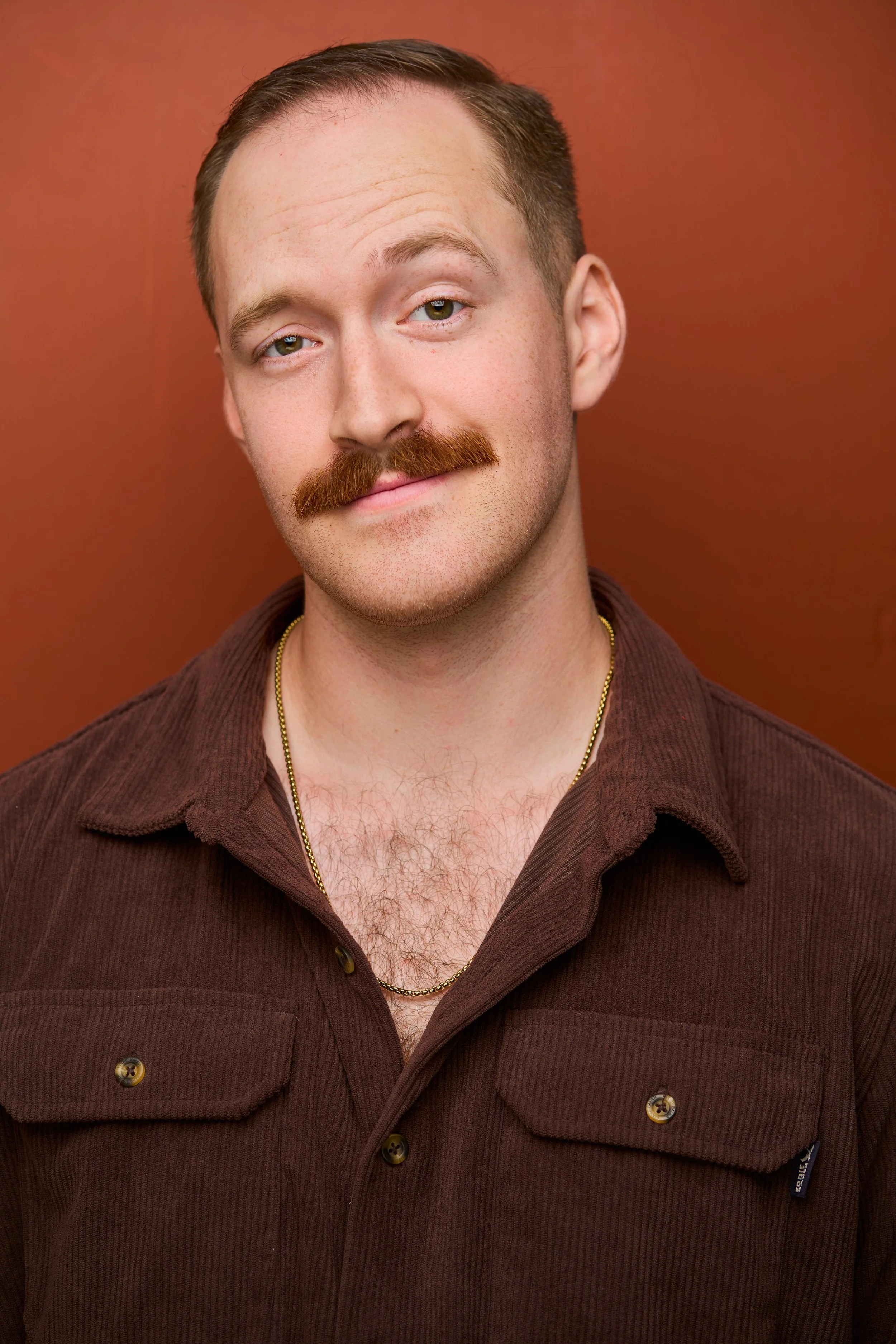 Close-up of a man with brown hair, light skin, a mustache, wearing a brown shirt, a gold chain, and standing against a brown background.