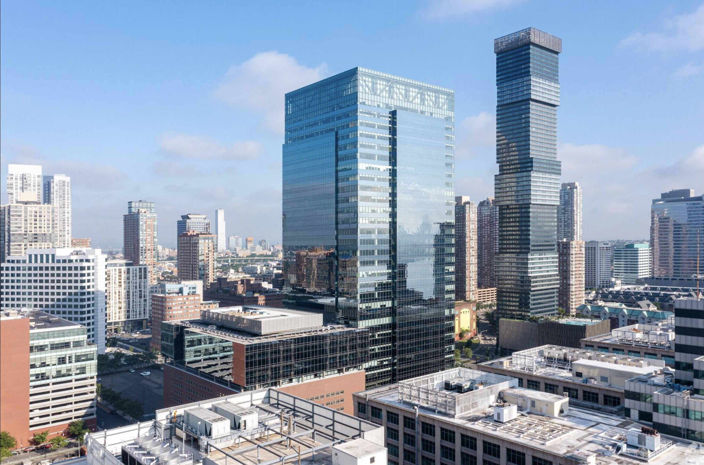 A city skyline featuring modern glass skyscrapers against a partly cloudy blue sky, with various commercial and residential buildings in the foreground.