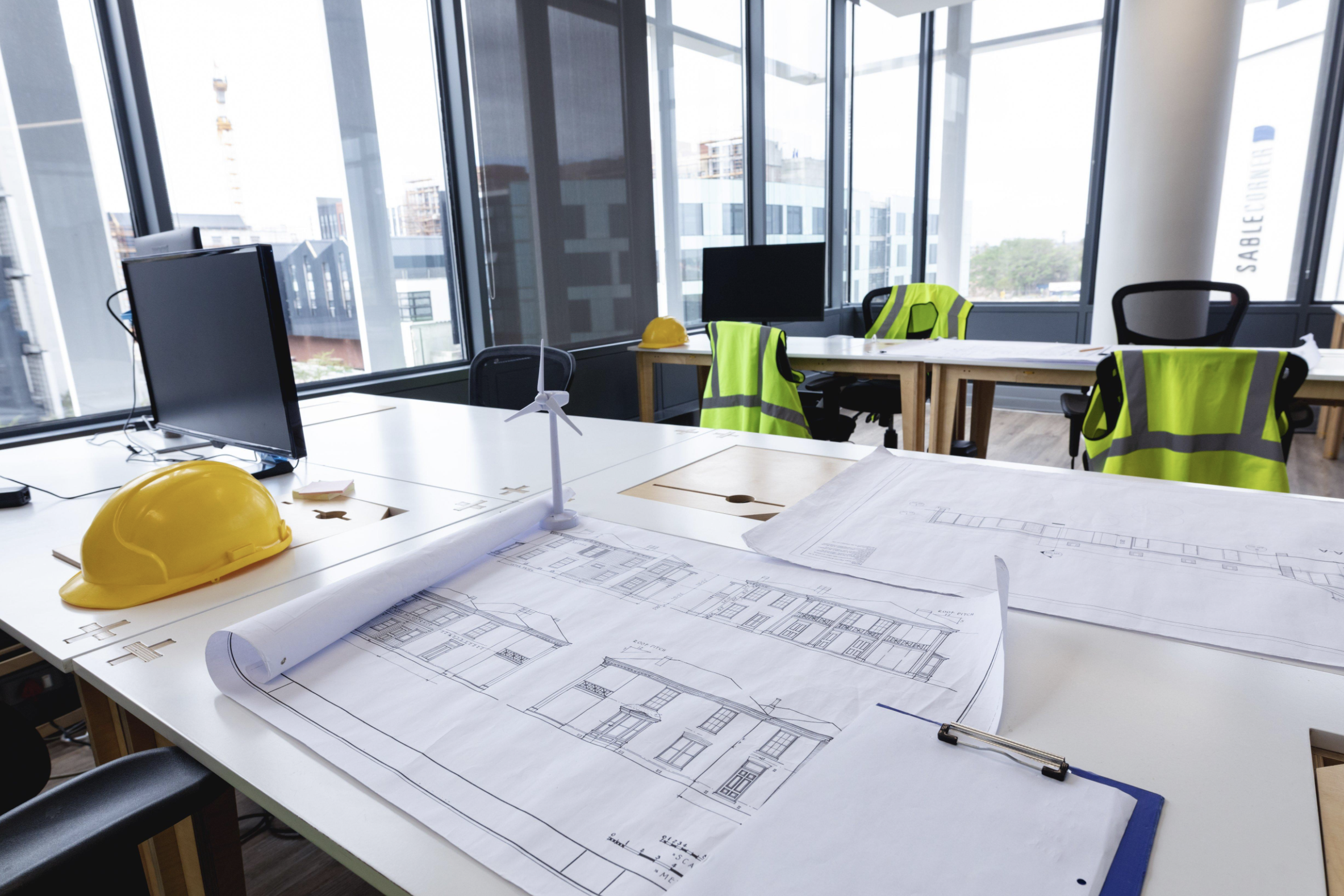 An office space with large windows showing city buildings outside. The desk has architectural blueprints, a yellow safety helmet, a small wind turbine model, and a clipboard with papers. There are yellow safety vests and black office chairs.