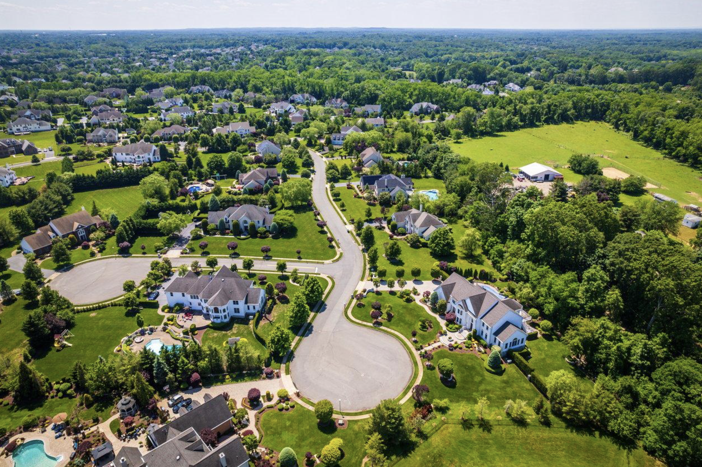 Aerial view of a suburban neighborhood with large homes, green lawns, trees, a park, and a large open green field surrounded by dense forest.