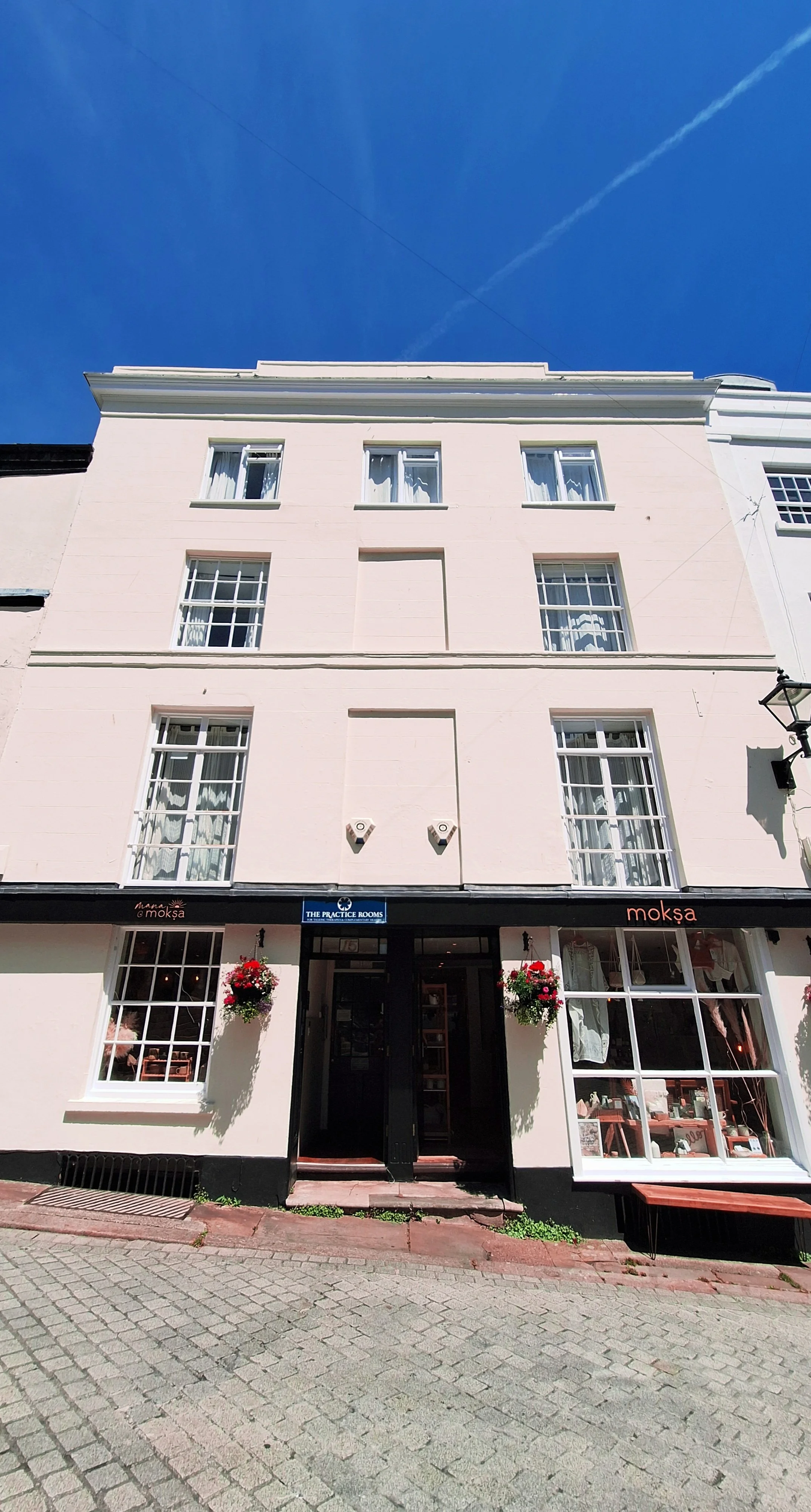 The Practice Rooms location. White multi-story building with large windows and black storefront on ground floor, featuring hanging flower baskets and a sign that reads 'The Practice Rooms'.