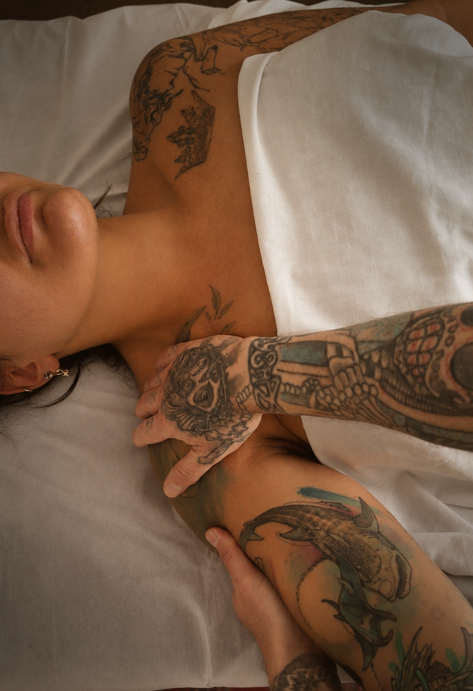 A person lying in bed with tattoos on their arms, chest, and neck, partly covered by a white sheet, with a neutral facial expression. Syluss performing massage.