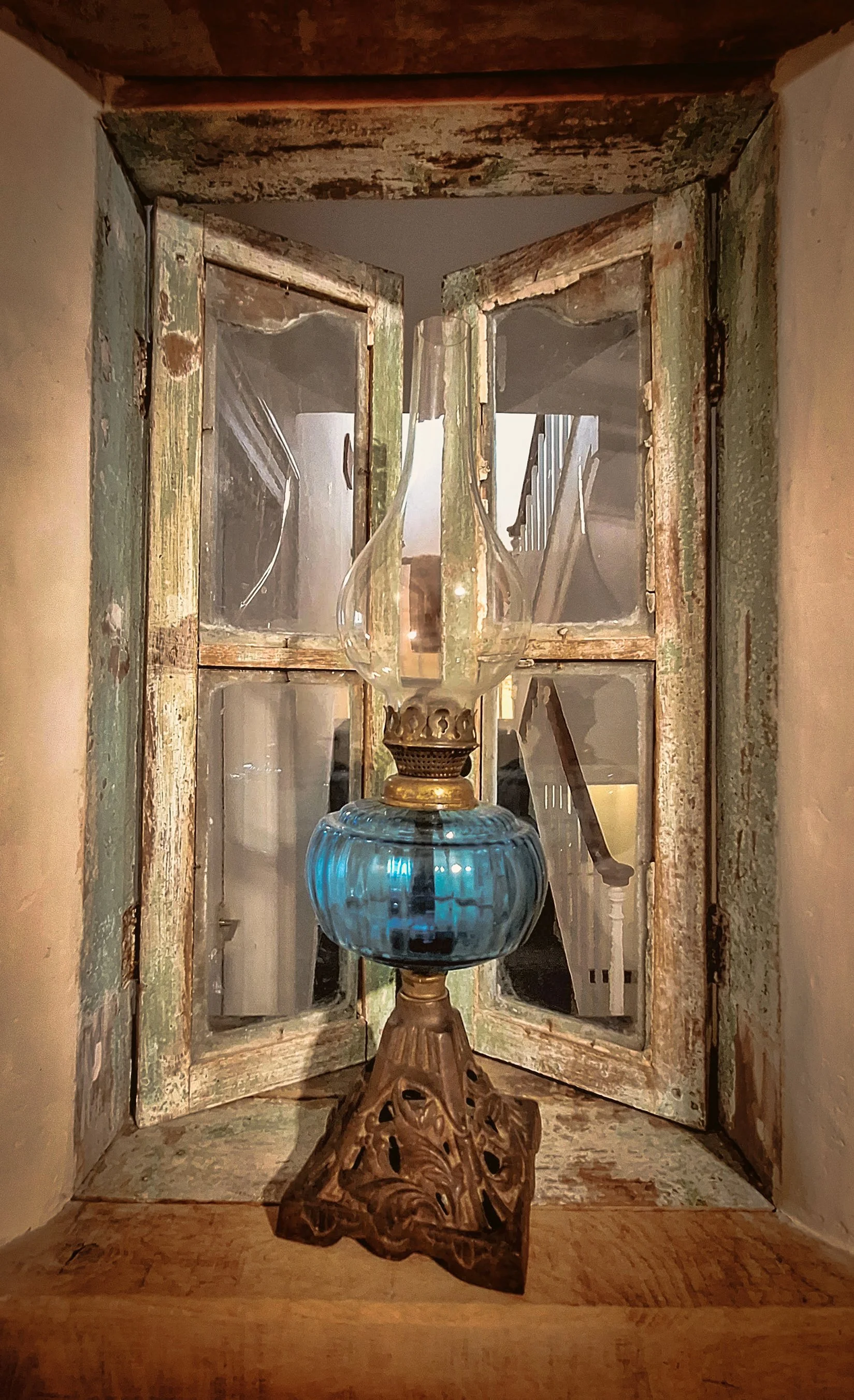 An antique oil lamp with a blue glass reservoir, intricate wooden base, and a glass chimney, placed in front of an old, distressed wooden window.