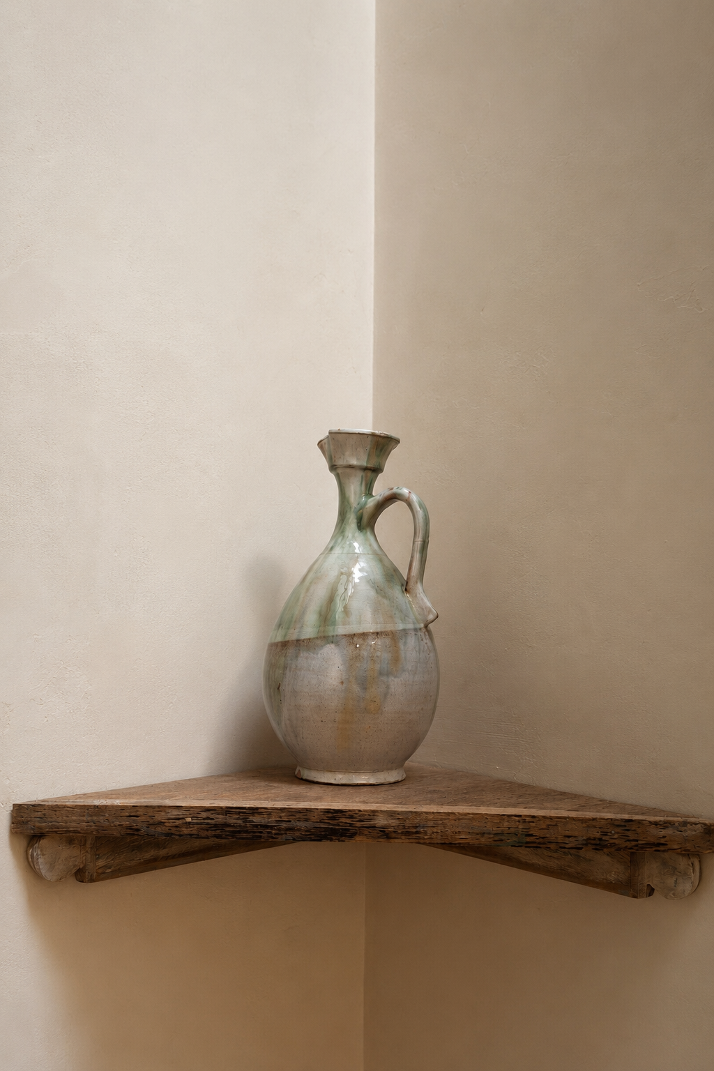 A ceramic vase with a handle and a narrow neck placed on a wooden shelf against a plain wall.
