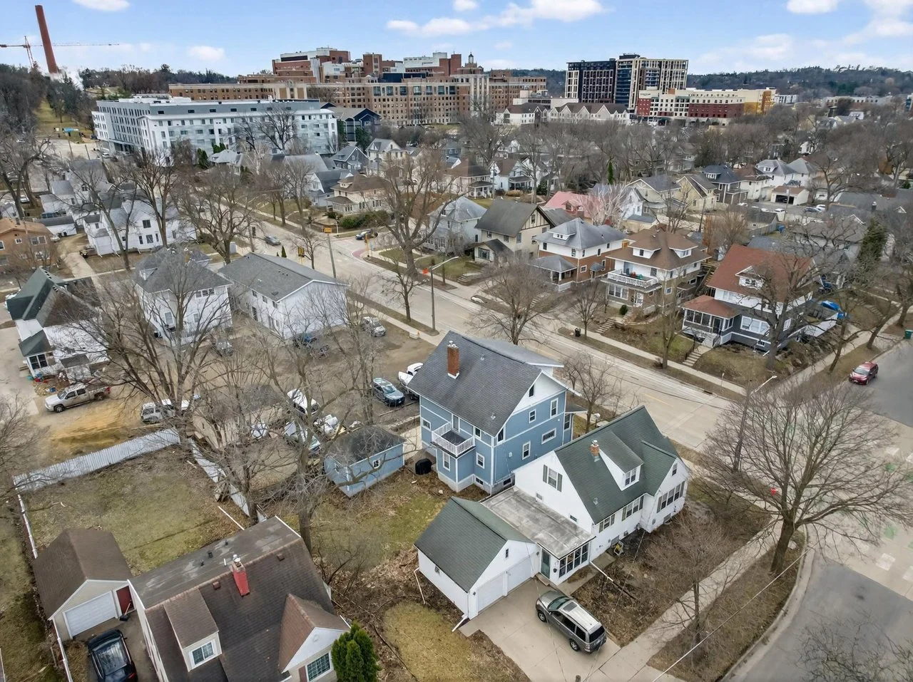 Aerial view of a residential neighborhood with single-family homes, trees, and cars parked along the streets, with a cityscape and taller buildings in the background.