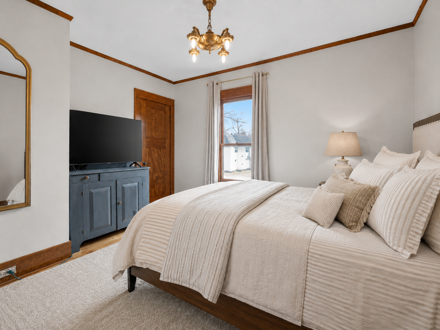 A cozy bedroom with a large bed, beige and striped bedding, beige pillows, a nightstand with a lamp, a window with white curtains, a blue TV cabinet with a flat-screen TV, a mirror on the wall, and a wooden door.