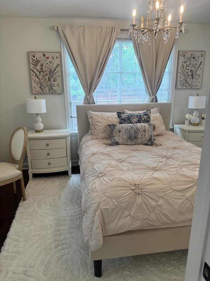 A cozy bedroom with a white upholstered bed, beige bedspread, and decorative pillows. Flanked by two white nightstands with lamps. Behind the bed, there are two large windows with beige curtains. A chandelier hangs from the ceiling, and framed botanical art decorates the wall.