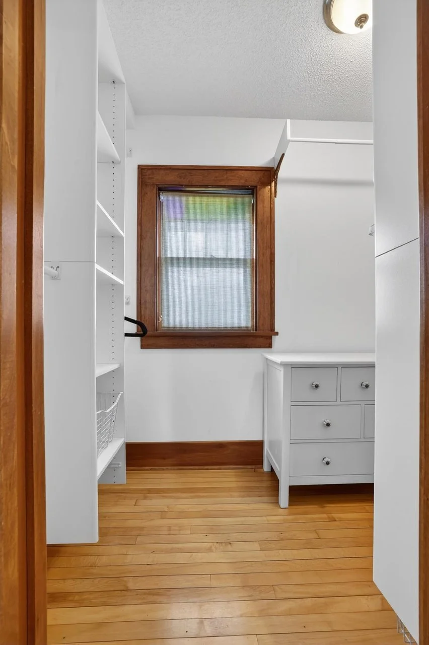 Empty laundry or utility room with a window, white built-in shelves, and a small white cabinet with drawers and knobs, with hardwood floors and white walls.