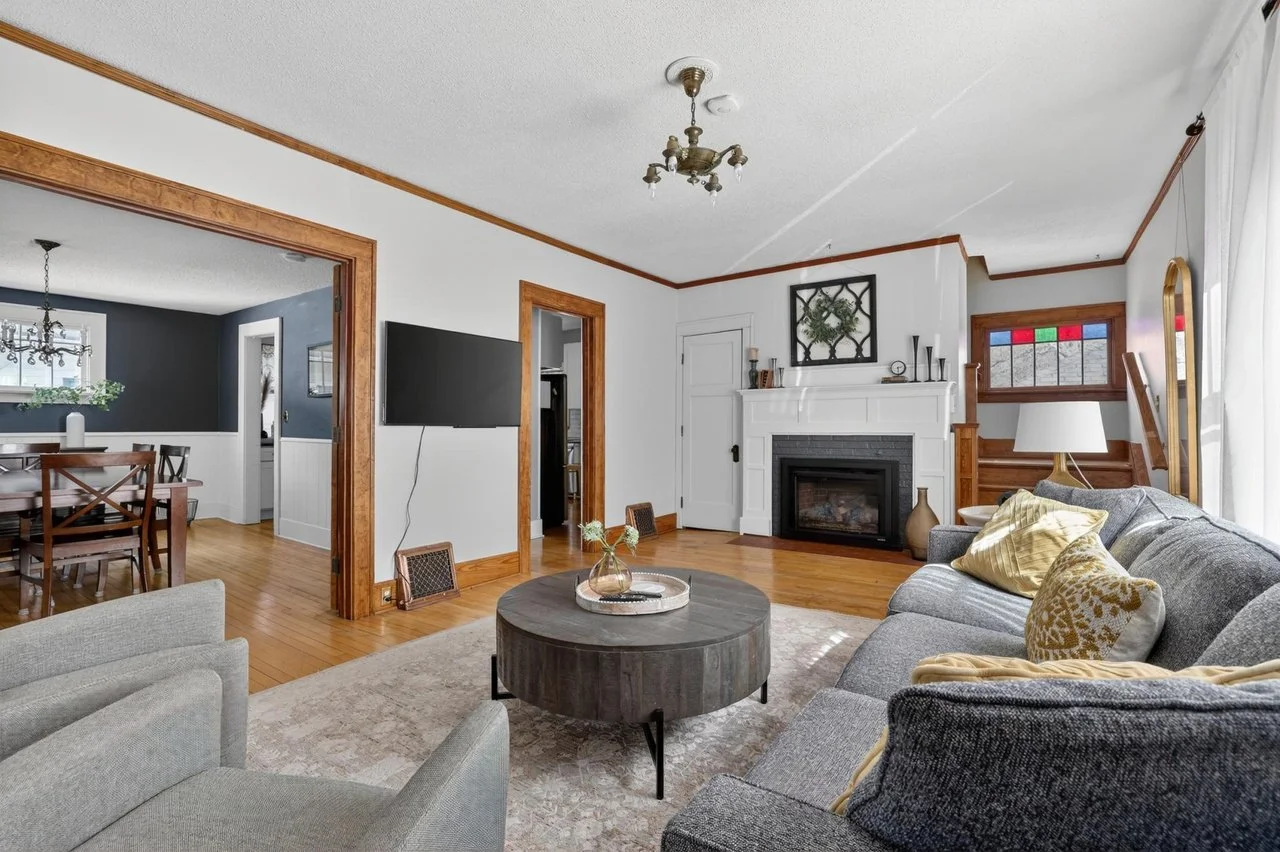 Living room with a gray sofa, yellow pillows, a dark round coffee table, a white fireplace, a wall-mounted TV, and a doorway leading to a patterned staircase.