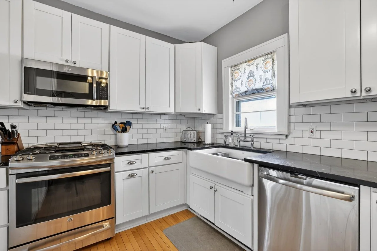A modern kitchen with white cabinets, black countertops, a stainless steel stove, microwave, dishwasher, and sink. There is a window with a patterned curtain above the sink, and wooden flooring.